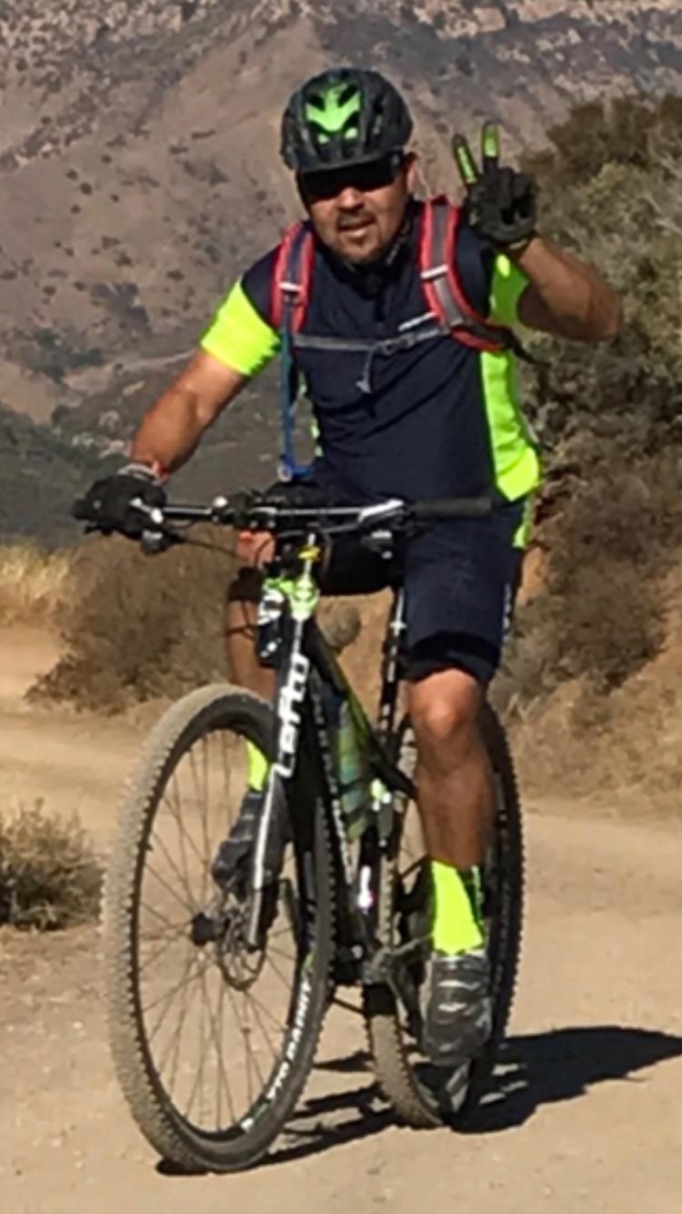 Cannondale F29 6: A mountain biker wearing a black helmet and bright green and black cycling attire rides along a dirt trail, giving a peace sign with his right hand. He is surrounded by a mountainous landscape.