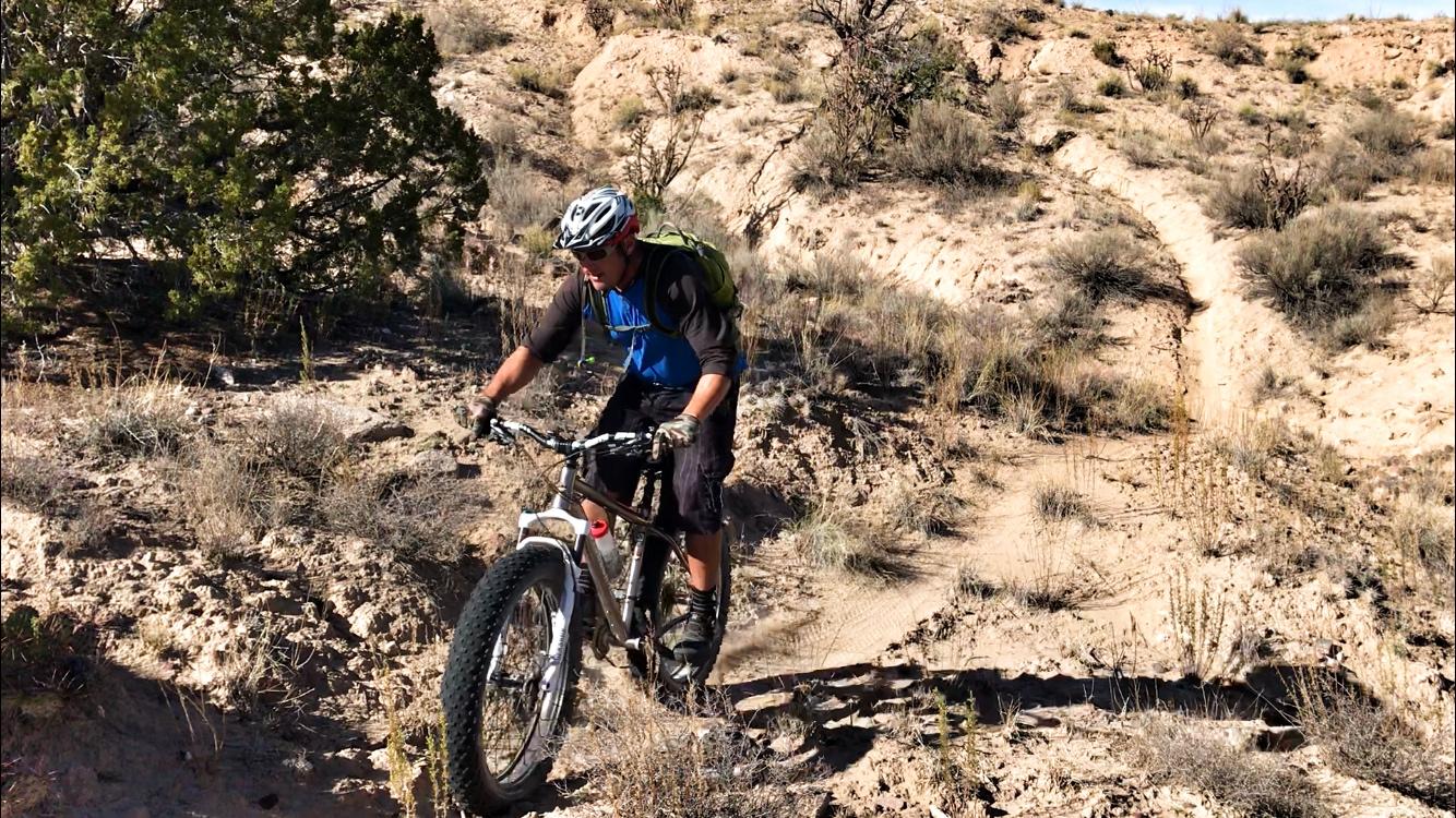 A mountain biker navigates a rugged dirt trail surrounded by low vegetation and rocky terrain. The rider, wearing a helmet and gloves, is focused on maintaining balance as they ride a fat-tire bicycle up a slope. The landscape features dry shrubs and sparse greenery typical of a desert environment. Mariposa Fat Bike Trails mountain bike trail.