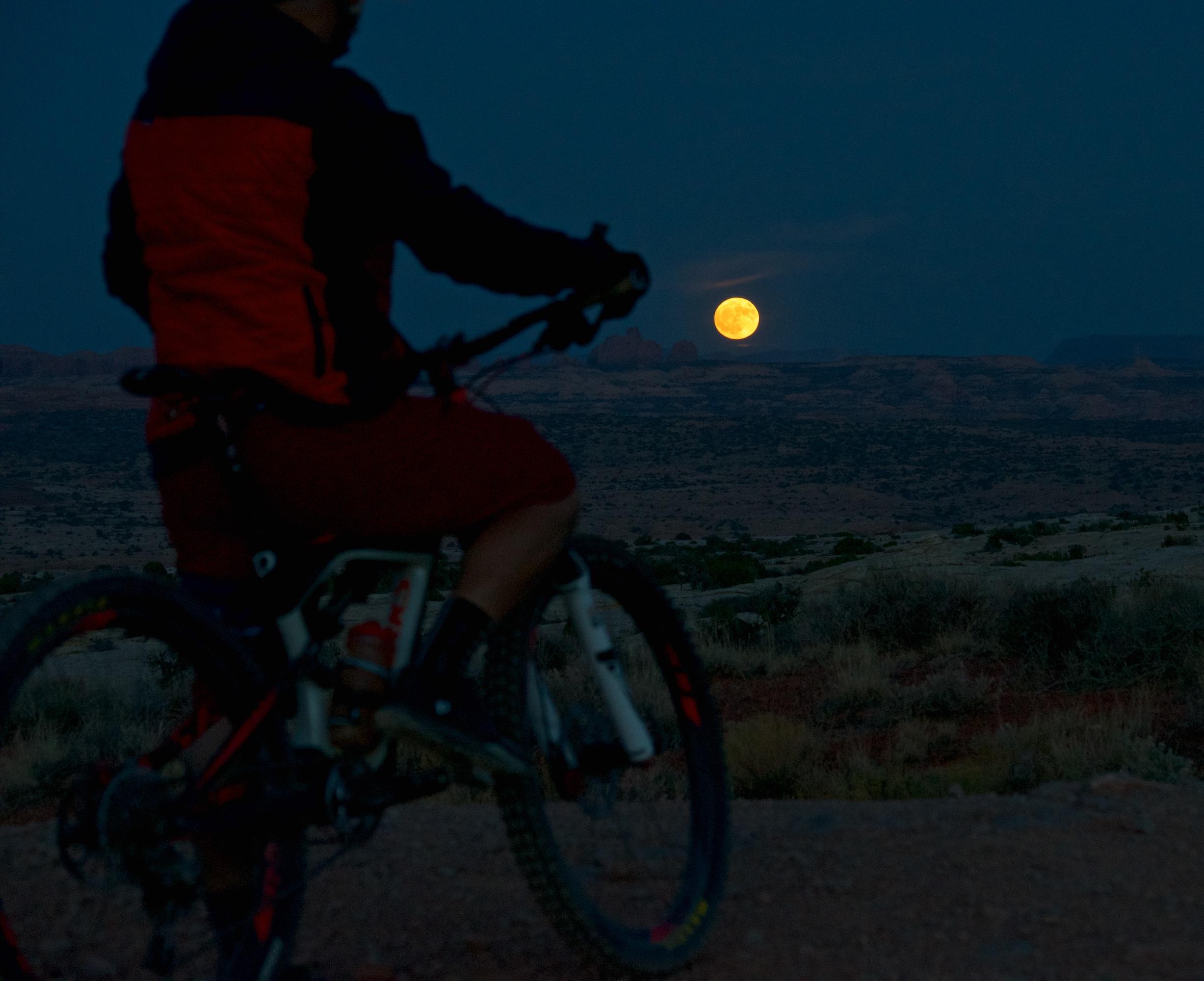 A cyclist in a red and black jacket sits on a mountain bike, silhouetted against a darkening sky as a large, bright full moon rises over a scenic landscape with rock formations and sparse vegetation. Moab Brand Trails mountain bike trail.