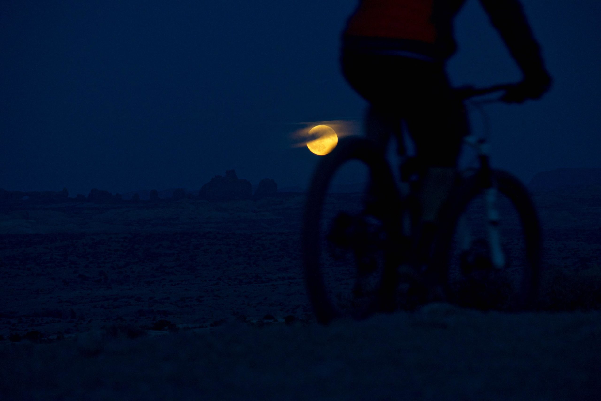 A silhouette of a cyclist in the foreground against a twilight sky, with a large moon partially obscured by clouds illuminating the landscape in the background. The scene captures a serene nighttime atmosphere in a rugged terrain. Moab Brand Trails mountain bike trail.
