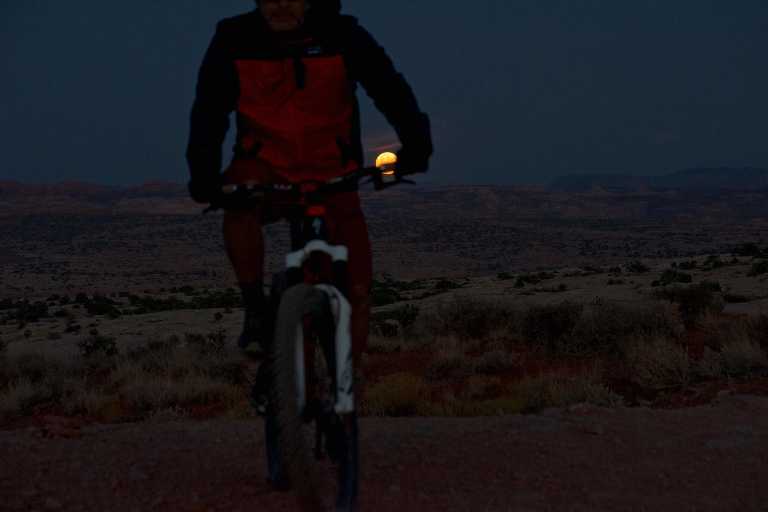 A cyclist rides on a dirt path under a night sky, with a partially visible moon rising in the background over a rocky landscape. The cyclist is wearing a red jacket, and their silhouette is highlighted against the darkening sky. Moab Brand Trails mountain bike trail.