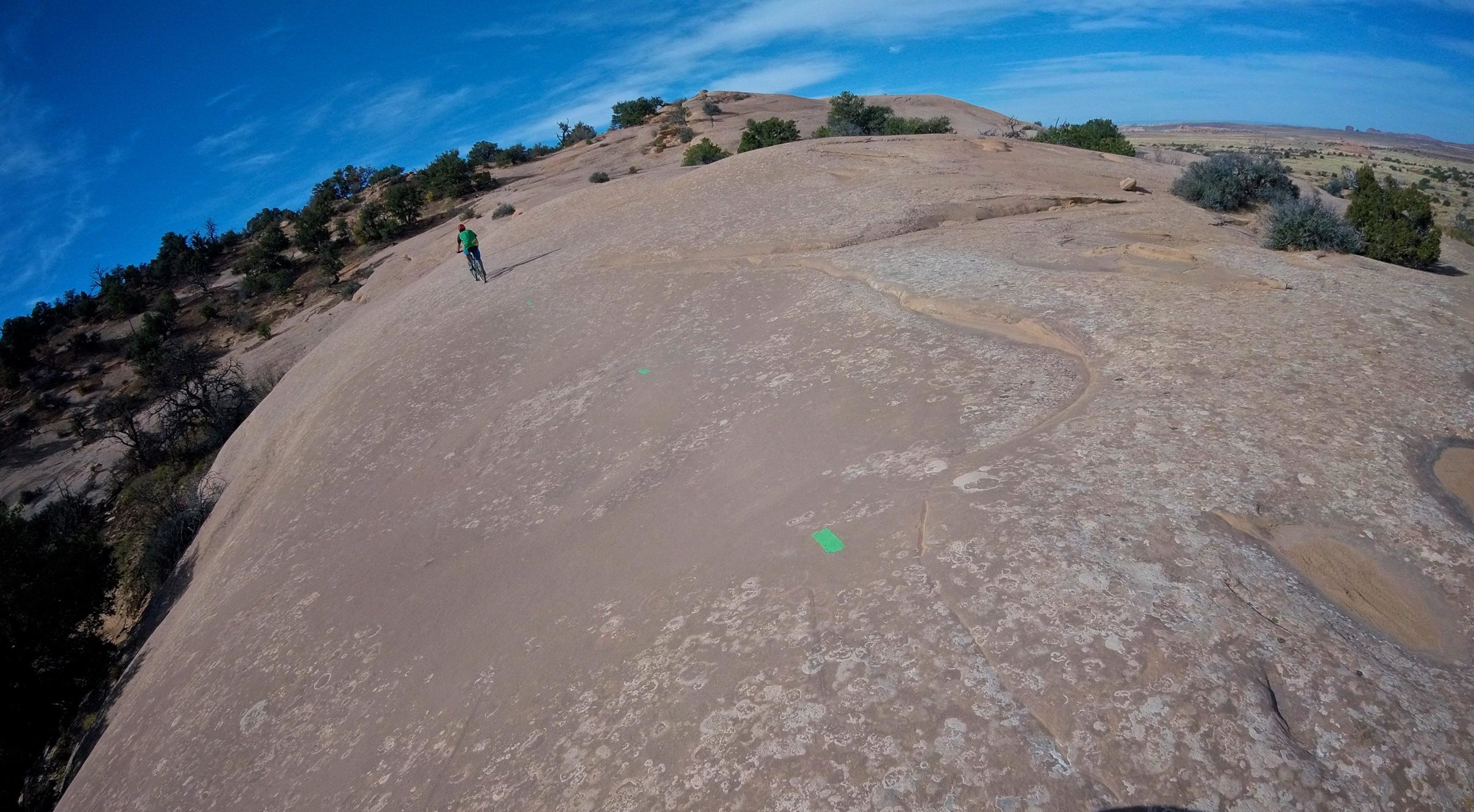 A person riding a bicycle along a rocky terrain, with a backdrop of a blue sky and distant hills. The surface is mostly smooth and features some green markers, indicating a trail, amidst sparse vegetation. Big Lonely mountain bike trail.