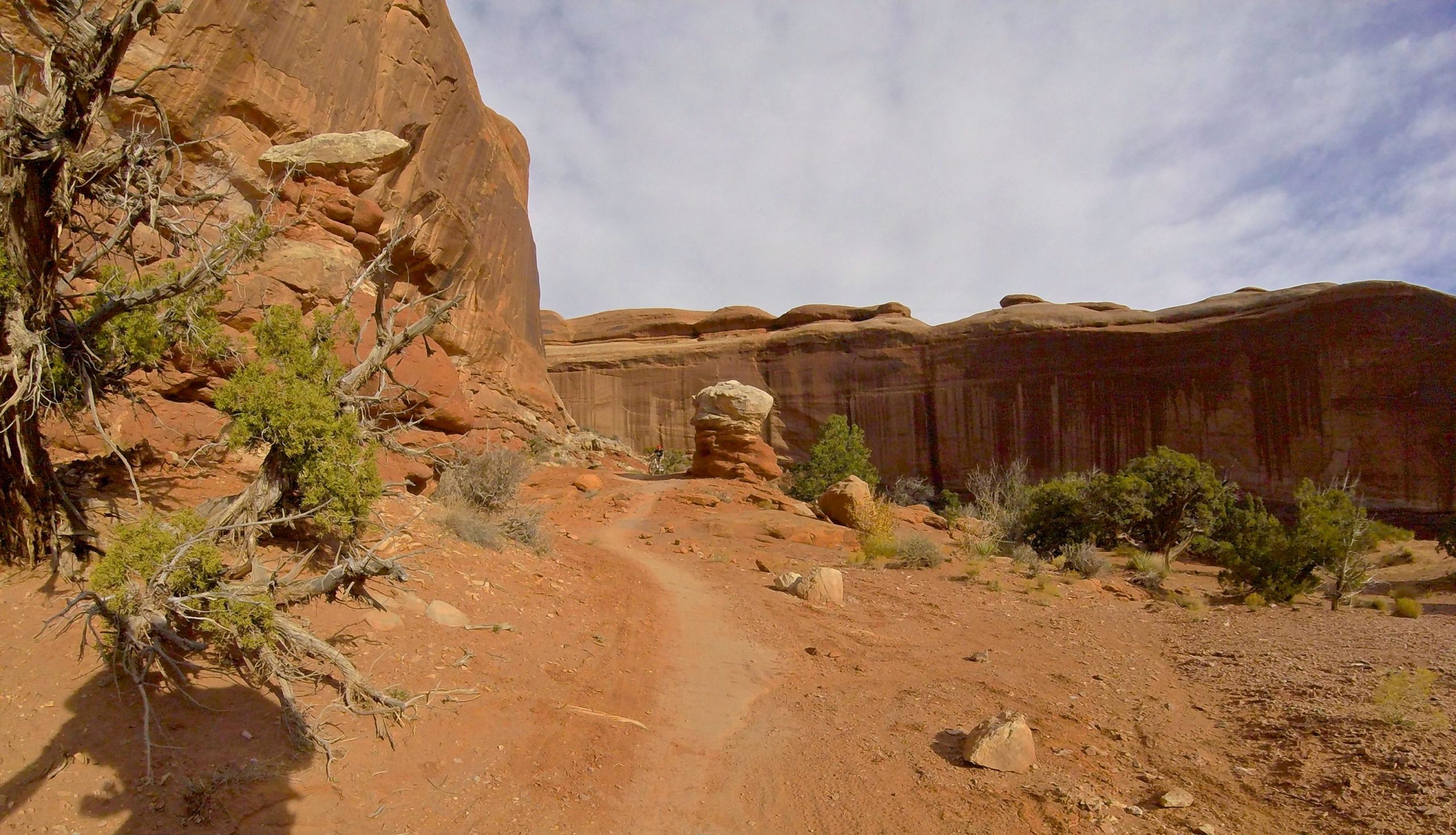 A winding dirt path leading through a rocky desert landscape, flanked by towering red rock formations and scattered vegetation. A large, distinctively shaped rock formation stands prominently along the path, under a partly cloudy sky. Big Mesa mountain bike trail.