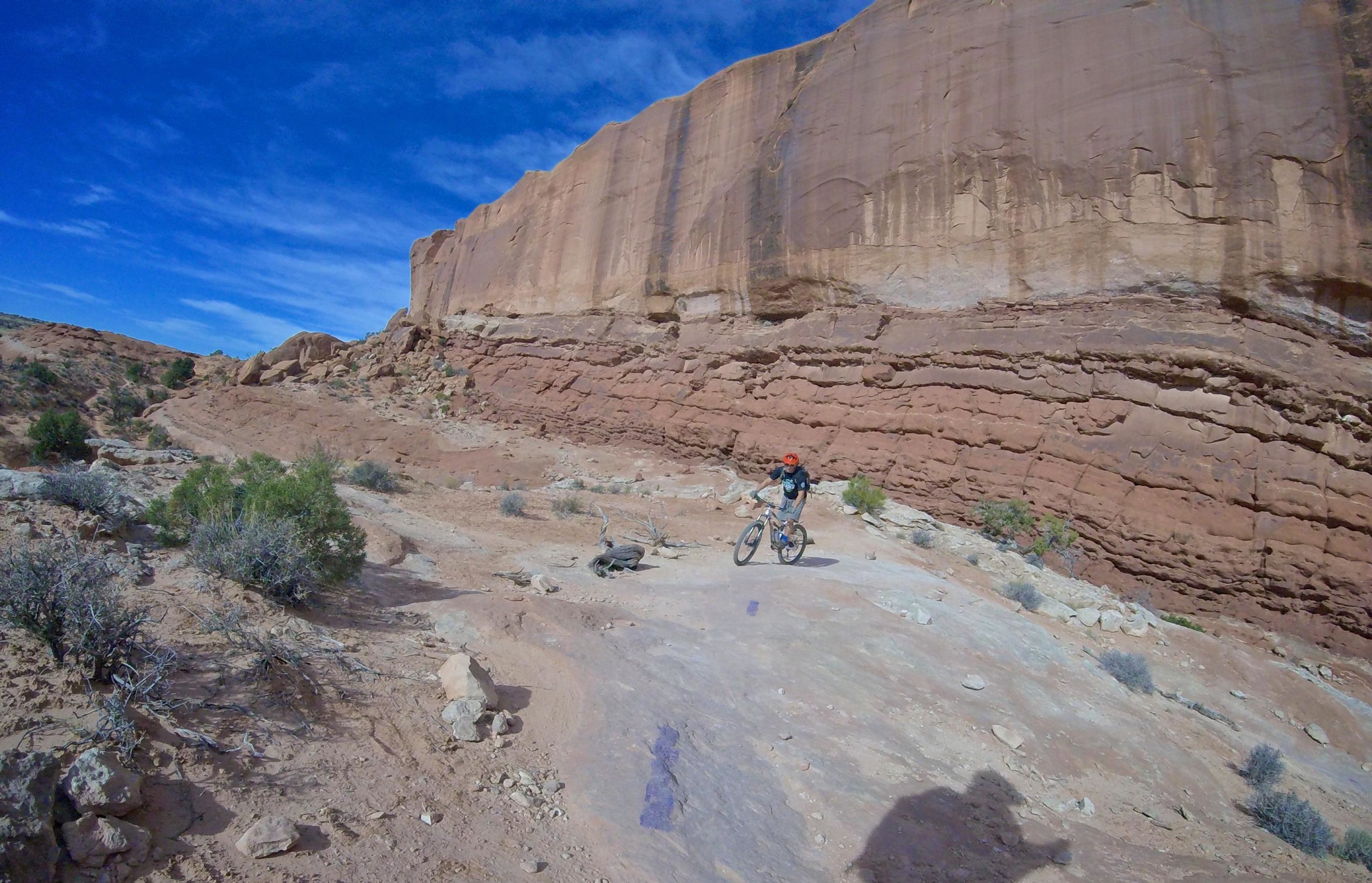 A mountain biker navigates a rocky trail surrounded by towering red rock formations under a bright blue sky. Vegetation is sparse, with small shrubs dotting the sandy landscape. Big Mesa mountain bike trail.