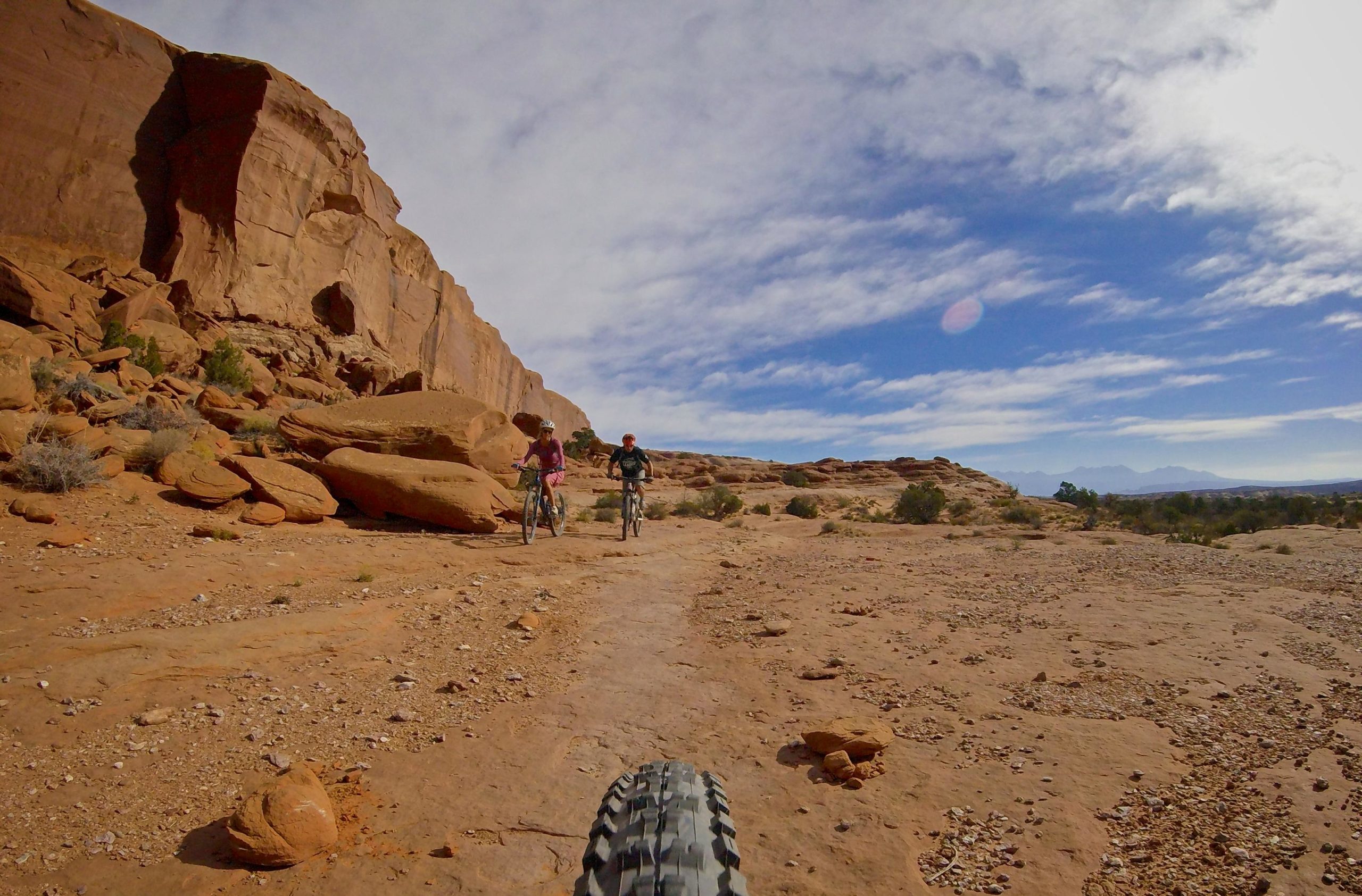 Two mountain bikers navigating a rocky trail in a desert landscape, surrounded by large rock formations under a partly cloudy sky. The view captures the perspective from a bike's front tire. Big Mesa mountain bike trail.