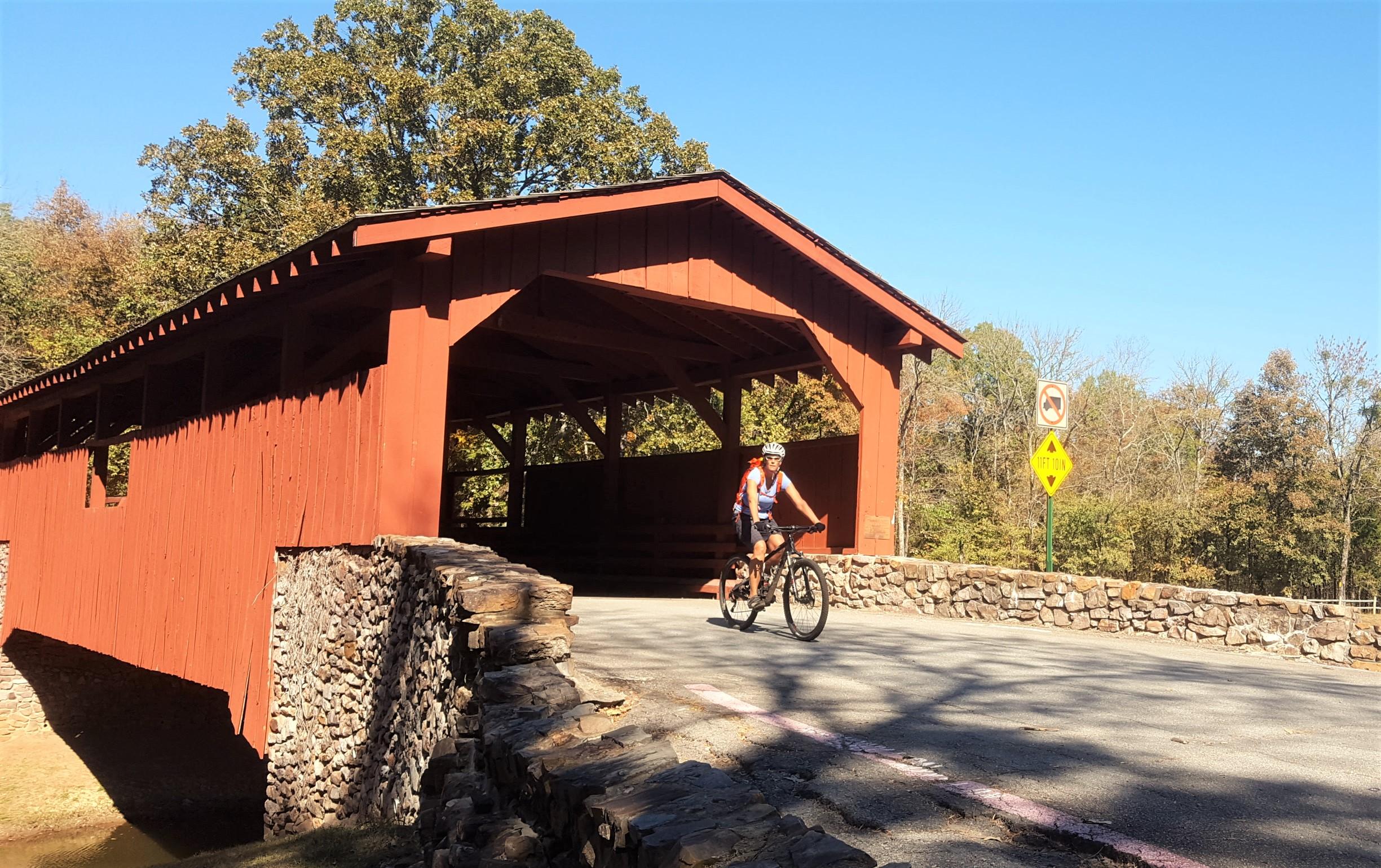 A cyclist riding a bike on a paved road near a red covered bridge with stone walls, surrounded by trees and a clear blue sky. Traffic signs are visible, indicating no left turn. The scene captures a sunny autumn day. Burns Park mountain bike trail.