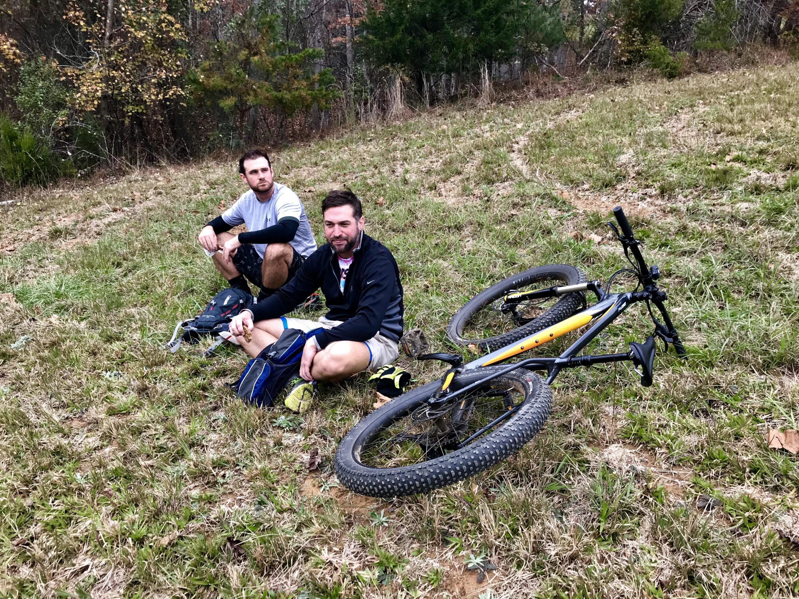 Two men sitting on the grass next to a mountain bike in a wooded area. One man is casually looking off to the side, while the other is facing the camera with a relaxed expression. Both are dressed in athletic clothing, and there are trees in the background. Warrior Creek mountain bike trail.