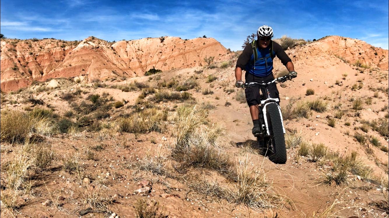 A person riding a fat tire mountain bike on a dusty trail in a rocky, desert landscape, with red rock formations and sparse vegetation in the background under a clear blue sky. Mariposa Fat Bike Trails mountain bike trail.
