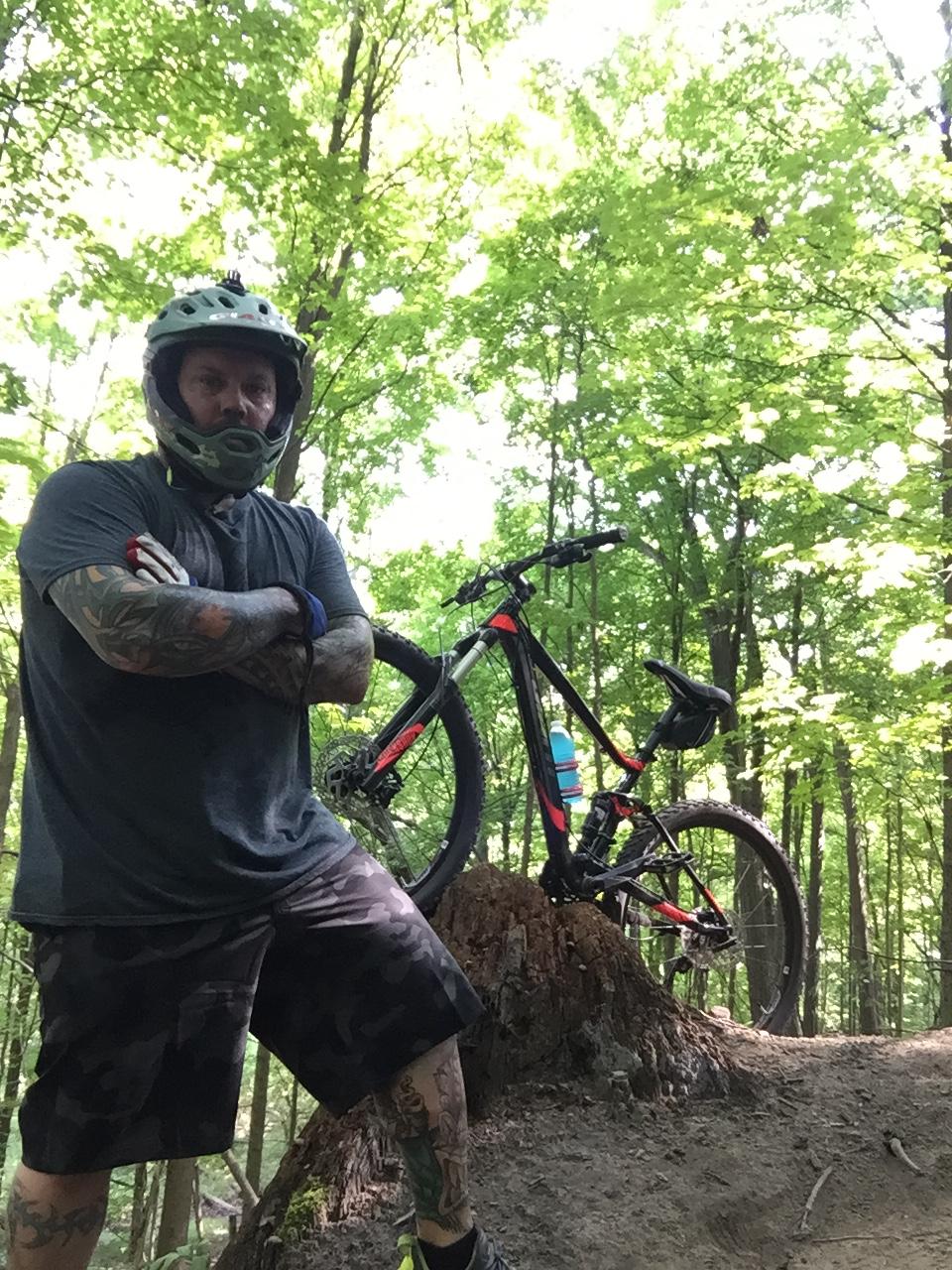 Giant Stance 27.5 2: A mountain biker in a helmet and protective gear stands confidently next to his bike, which is propped up on a large tree stump. The scene is set in a lush, green forest with sunlight filtering through the leaves, creating a vibrant outdoor atmosphere. The biker has tattoos visible on his arms and is dressed in casual biking attire.