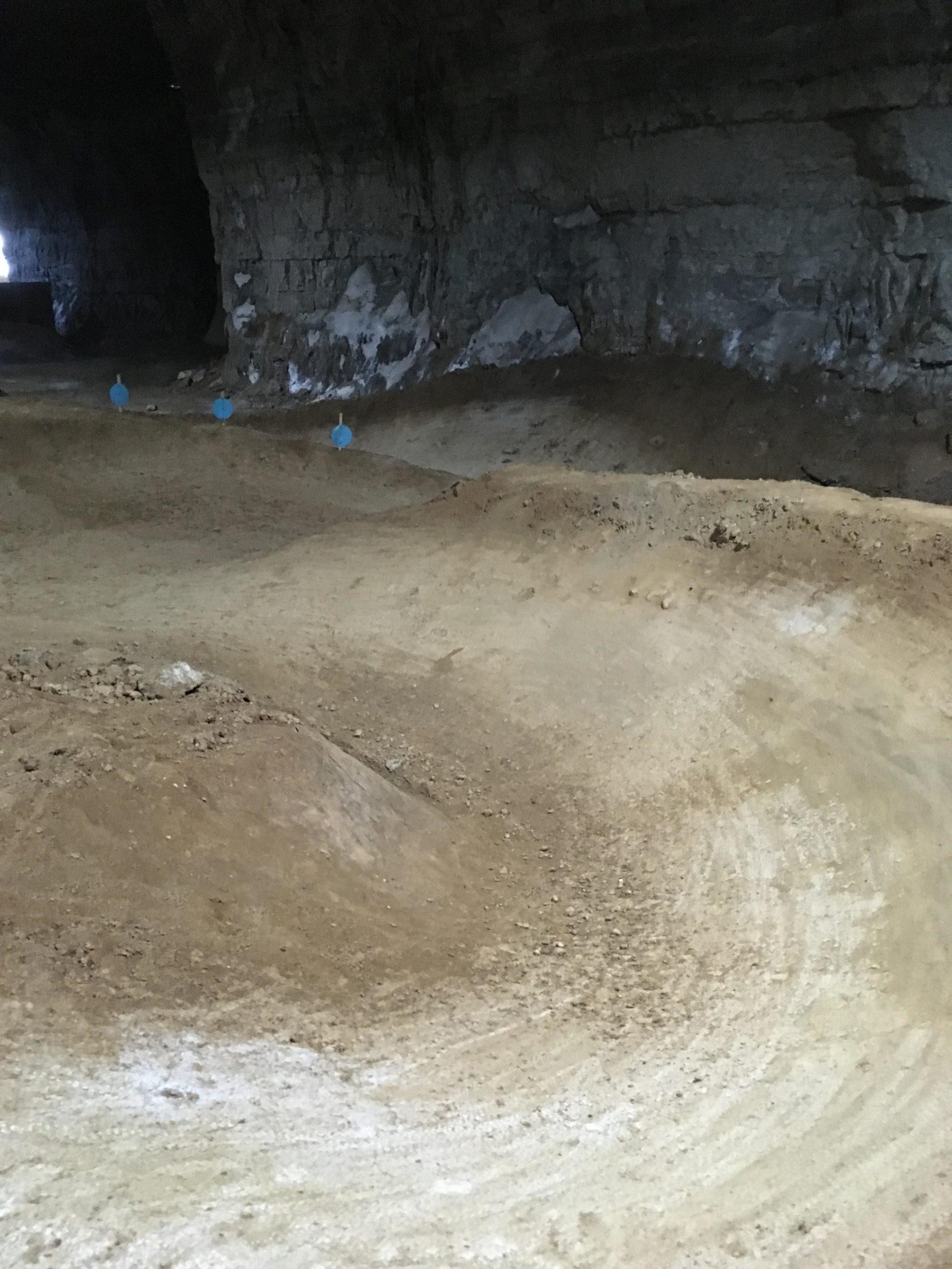 A dimly lit underground space with sandy terrain featuring several dirt mounds and curves, hinting at a potential biking or ATV track. In the background, a rocky wall and faint light can be seen. Blue markers are positioned along the track. Louisville Mega Cavern Bike Park mountain bike trail.