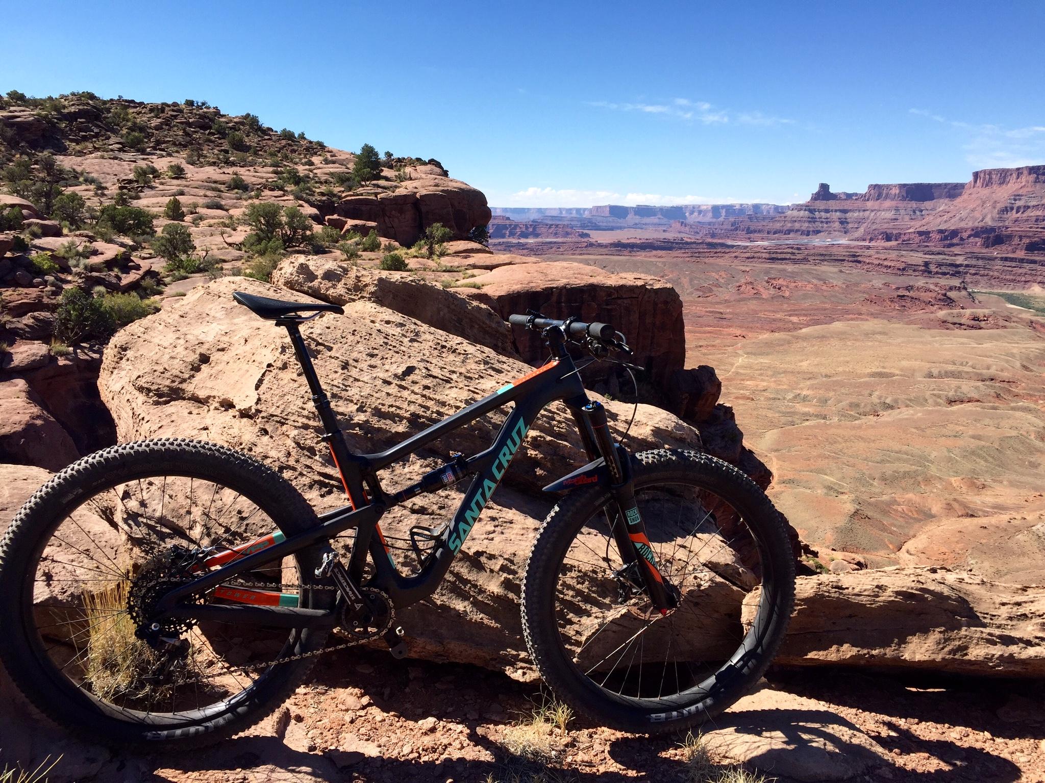 Santa Cruz Hightower 29er/27.5: A mountain bike resting on a rocky outcrop, overlooking a vast canyon landscape with layered red rock formations under a clear blue sky.
