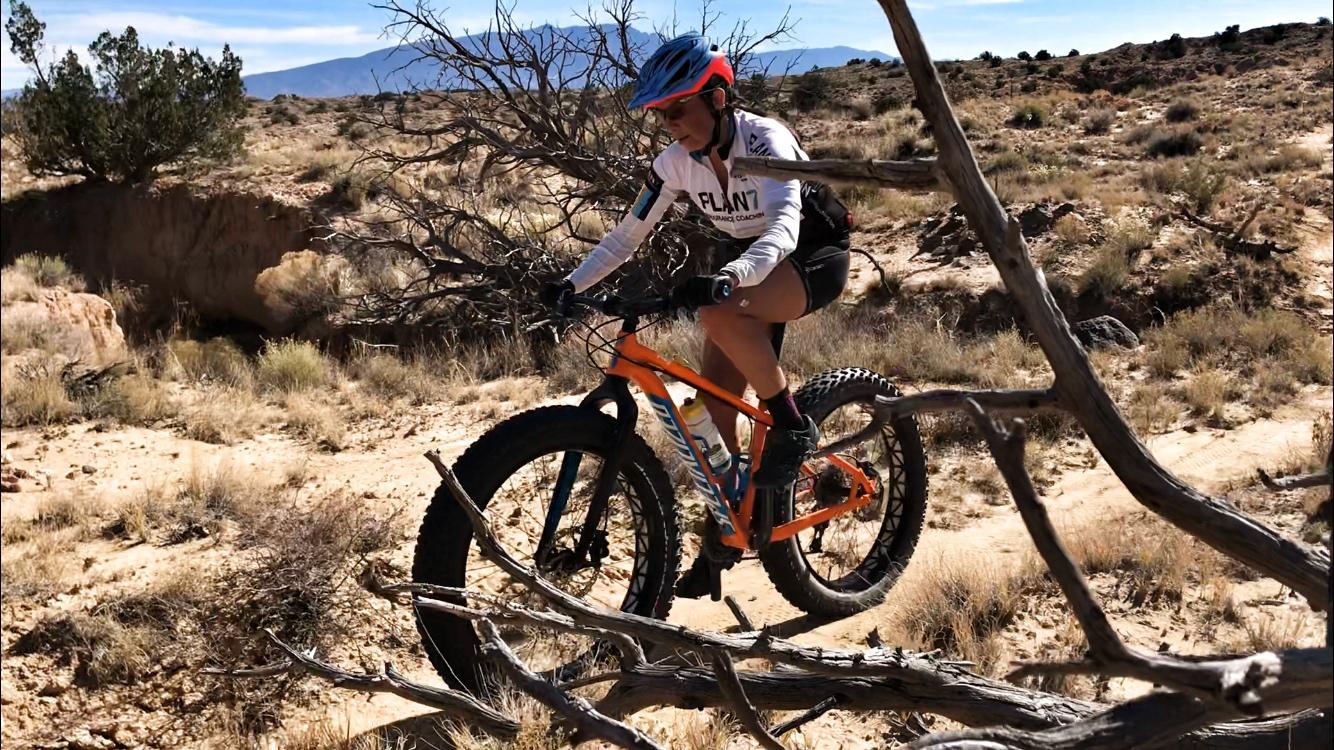 A person riding a mountain bike with large tires across a rugged terrain, navigating around a fallen branch. The background features dry shrubbery and distant mountains under a clear blue sky. Mariposa Fat Bike Trails mountain bike trail.