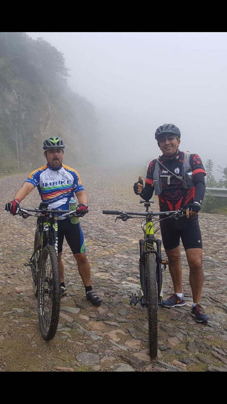 Cannondale F29 6: Two mountain bikers pose on a foggy, rocky road. One cyclist, wearing a blue and yellow jersey, holds a water bottle and looks toward the camera. The other, dressed in a red and black shirt, gives a thumbs-up. Surrounding them is a misty landscape, adding to the adventurous atmosphere of the scene.