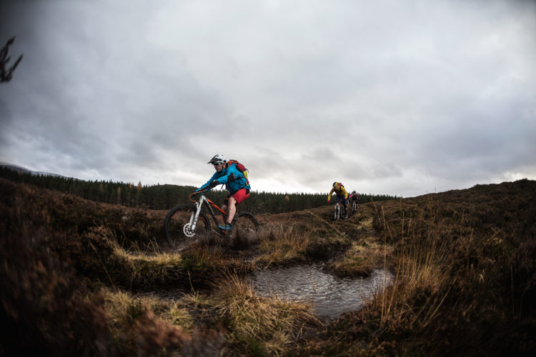 Two mountain bikers navigate a rugged trail through heather-covered terrain, with patches of standing water and a cloudy sky overhead. The cyclists are wearing helmets and colorful jackets, while a forest is visible in the background.