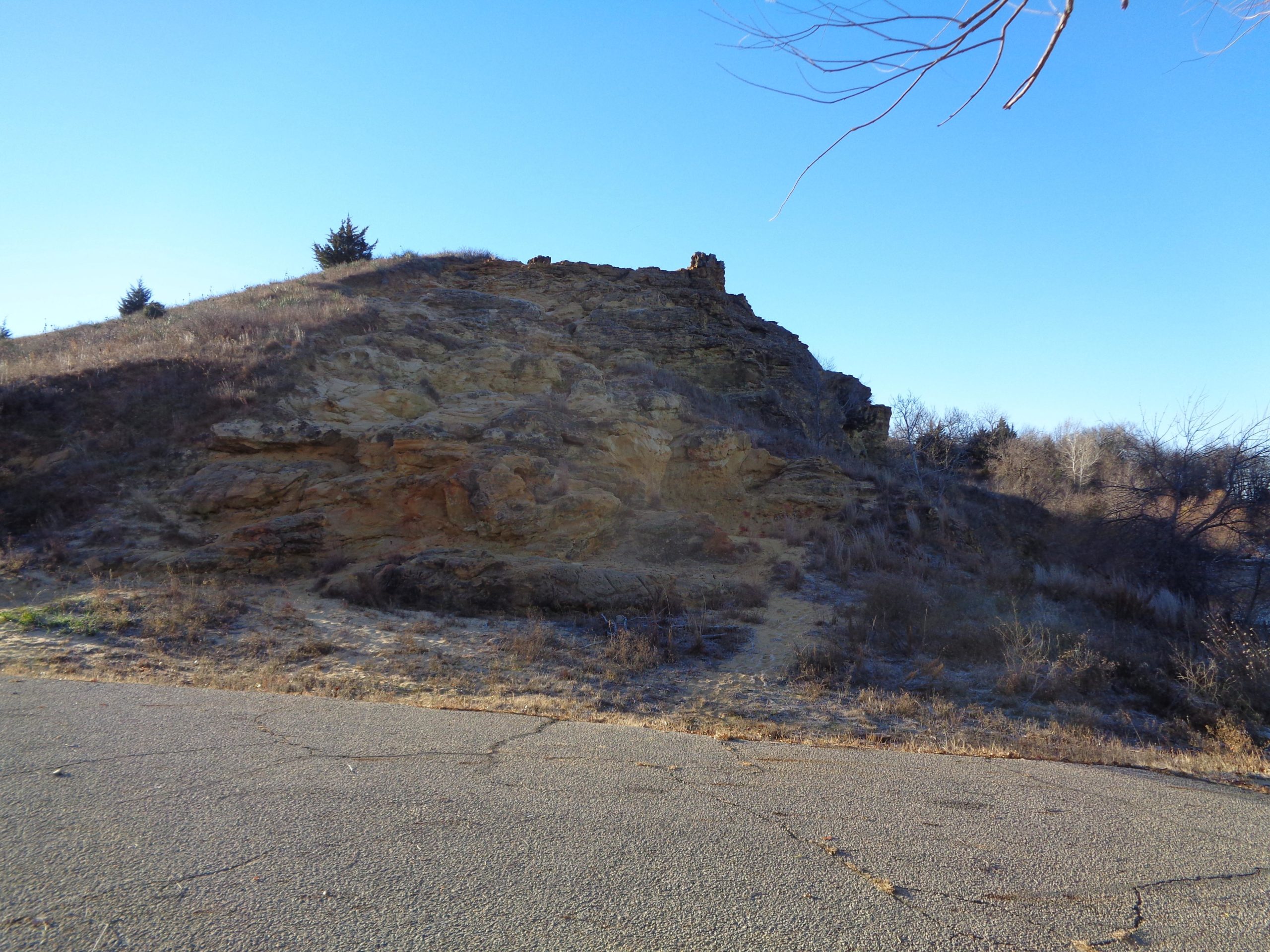 A rocky hill with sparse vegetation, surrounded by a clear blue sky. The foreground shows a cracked asphalt surface leading up to the hill, while small trees and shrubs are visible on and around the slope. Kanopolis Lake mountain bike trail.