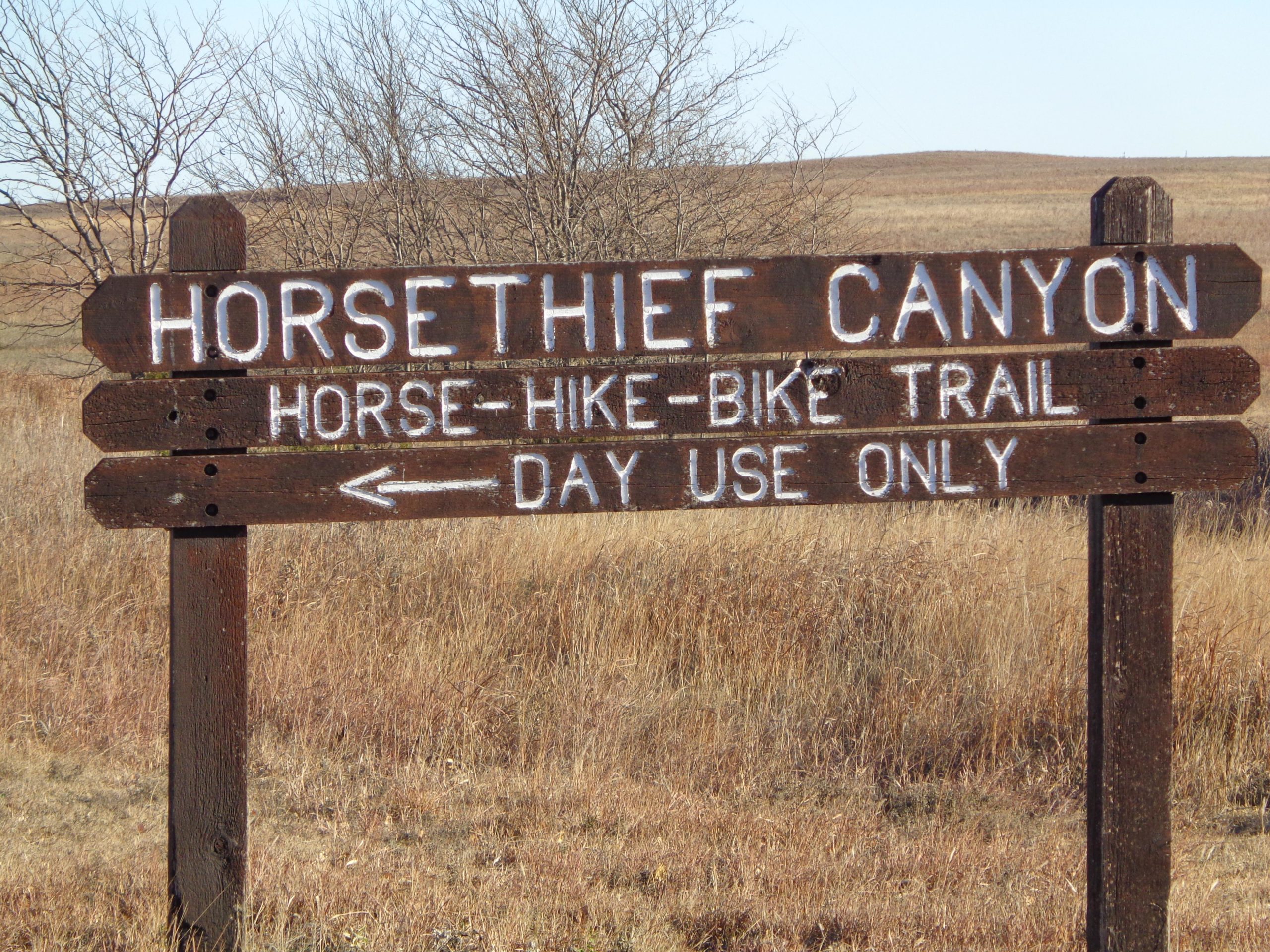 A wooden trail sign for Horsethief Canyon indicating a horse, hike, and bike trail, with an arrow pointing left and stating "Day Use Only." The sign is situated in a grassy area with sparse trees in the background. Kanopolis Lake mountain bike trail.