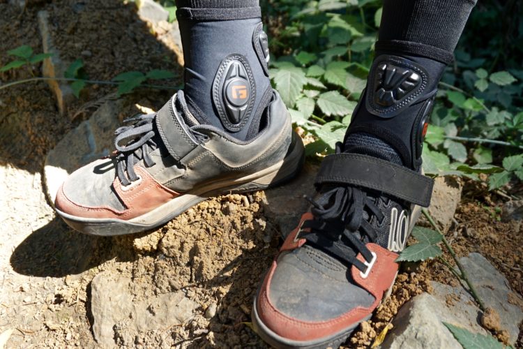 A close-up of two pairs of outdoor shoes resting on rocky ground, surrounded by greenery. One shoe is brown and grey, featuring a sturdy design with laces and a velcro strap, while the other is primarily black with red accents, showcasing a typical athletic style. Both shoes are worn and slightly dirty, indicating use in outdoor activities.