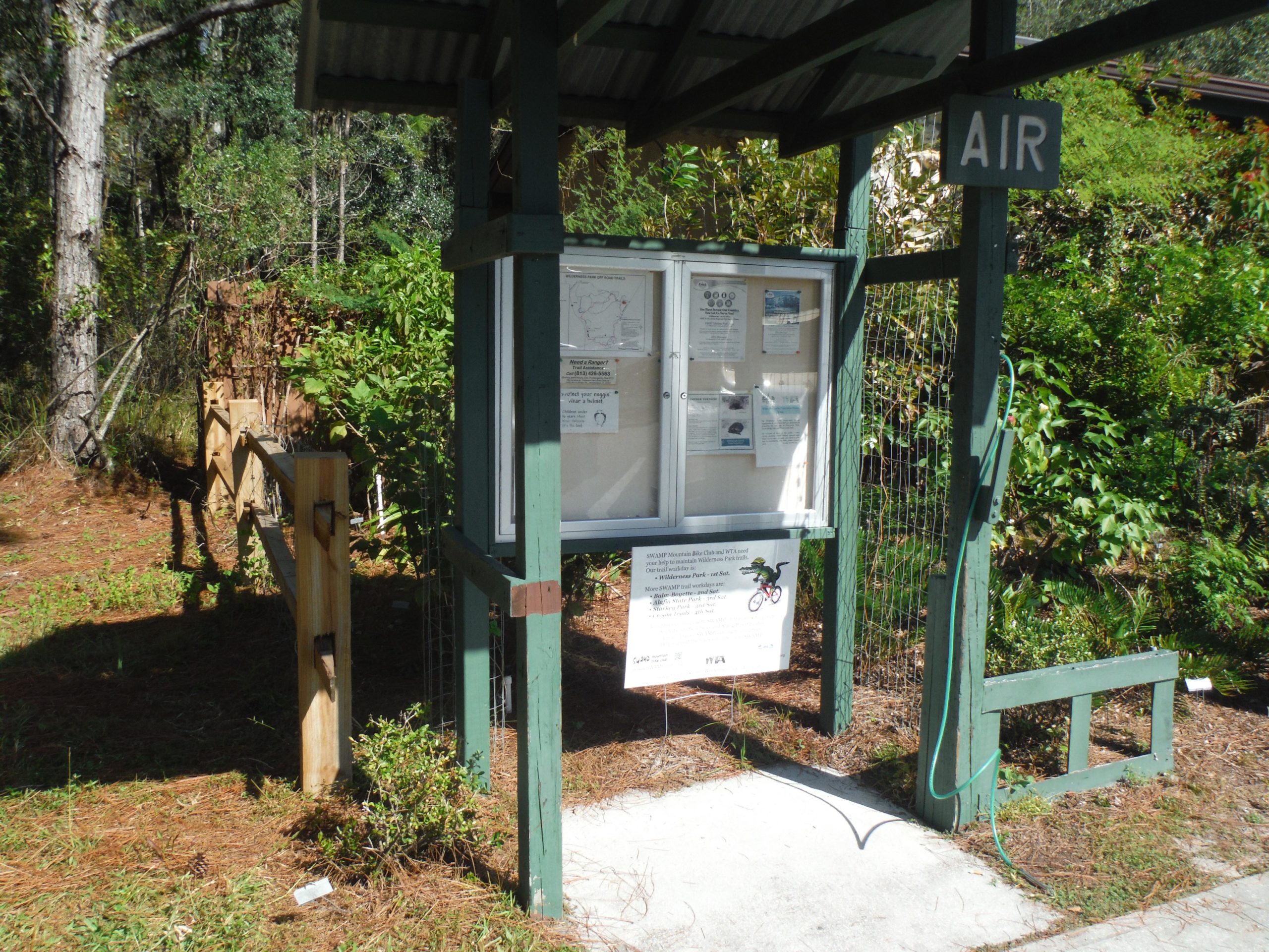 A small wooden structure with a roof, featuring a sign labeled "AIR." The front has a display board with various notices and maps. Surrounding the structure is lush greenery and pine needles on the ground, with a wooden fence visible on the left. Flatwoods Park mountain bike trail.