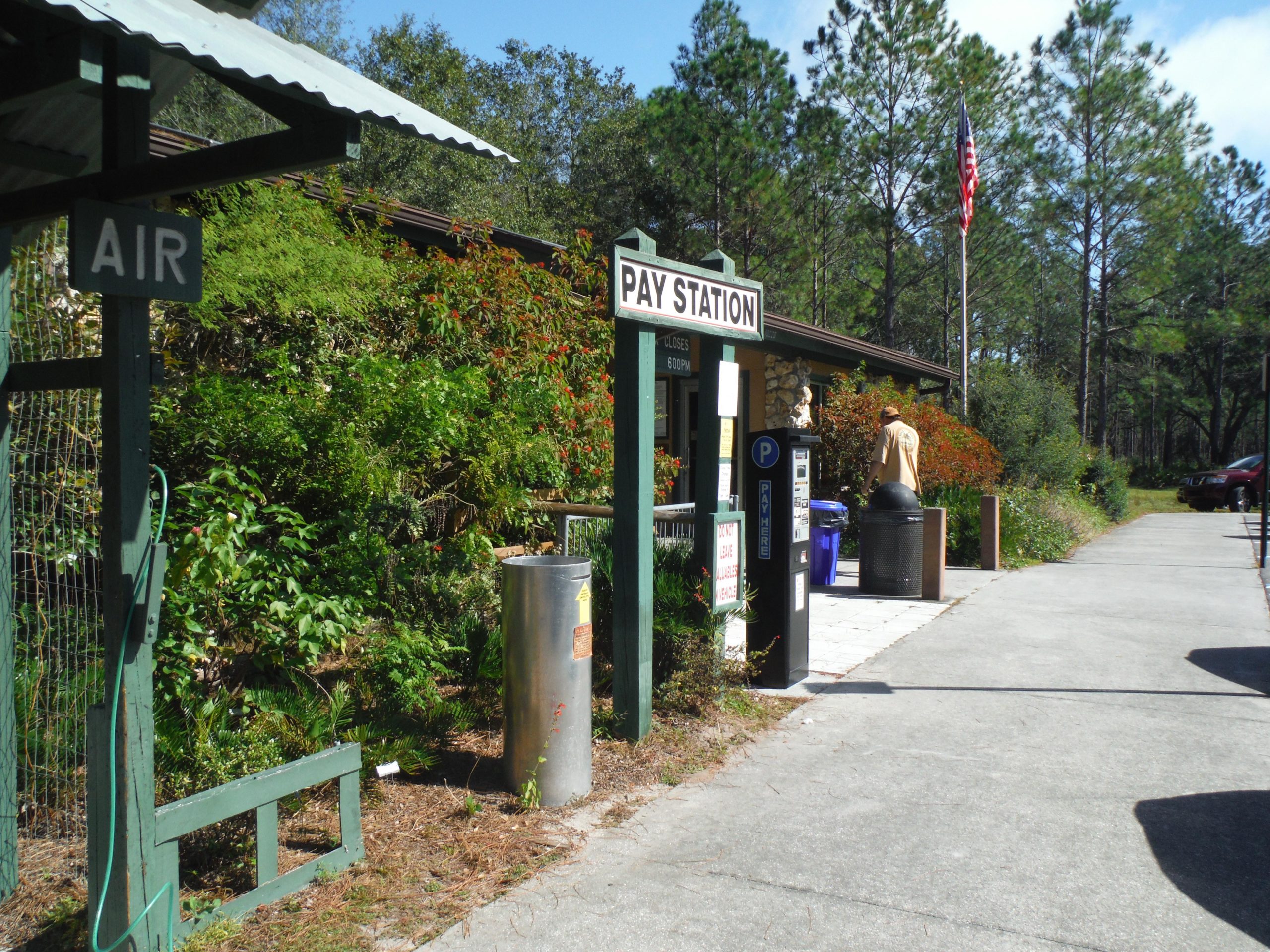 A view of a pay station in a natural setting, with a sign indicating "PAY STATION" and another sign labeled "AIR." Green foliage surrounds the area, and a pathway leads towards a building. A person in a tan shirt is visible near the pay station, and a flag can be seen in the background. Vegetation and parked cars are also present in the scene. Flatwoods Park mountain bike trail.