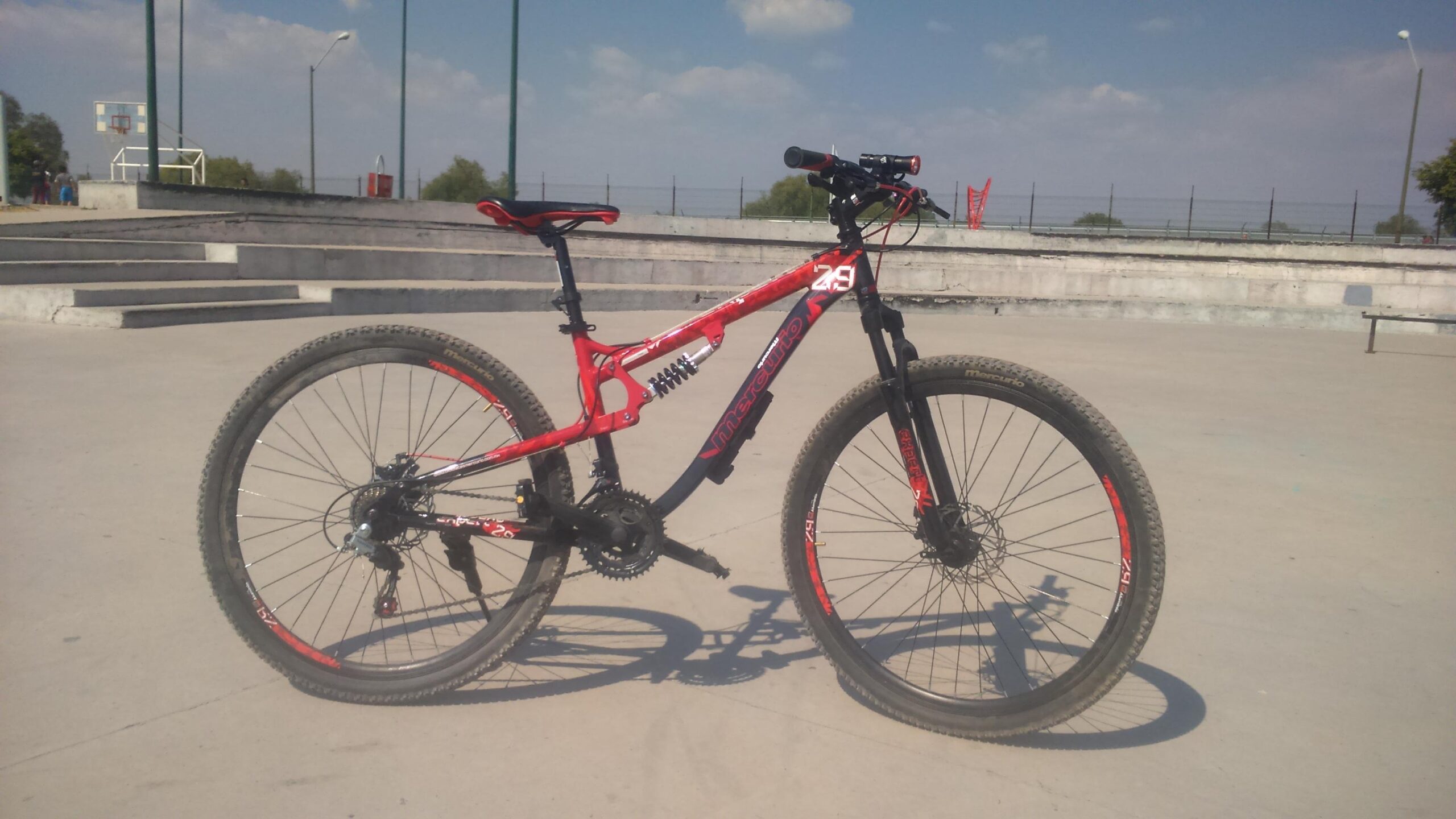 Mercurio Mercurio Expert: A red mountain bike with black accents is parked on a concrete surface, featuring a visible front suspension and knobby tires. The background includes a basketball court and clear blue skies.