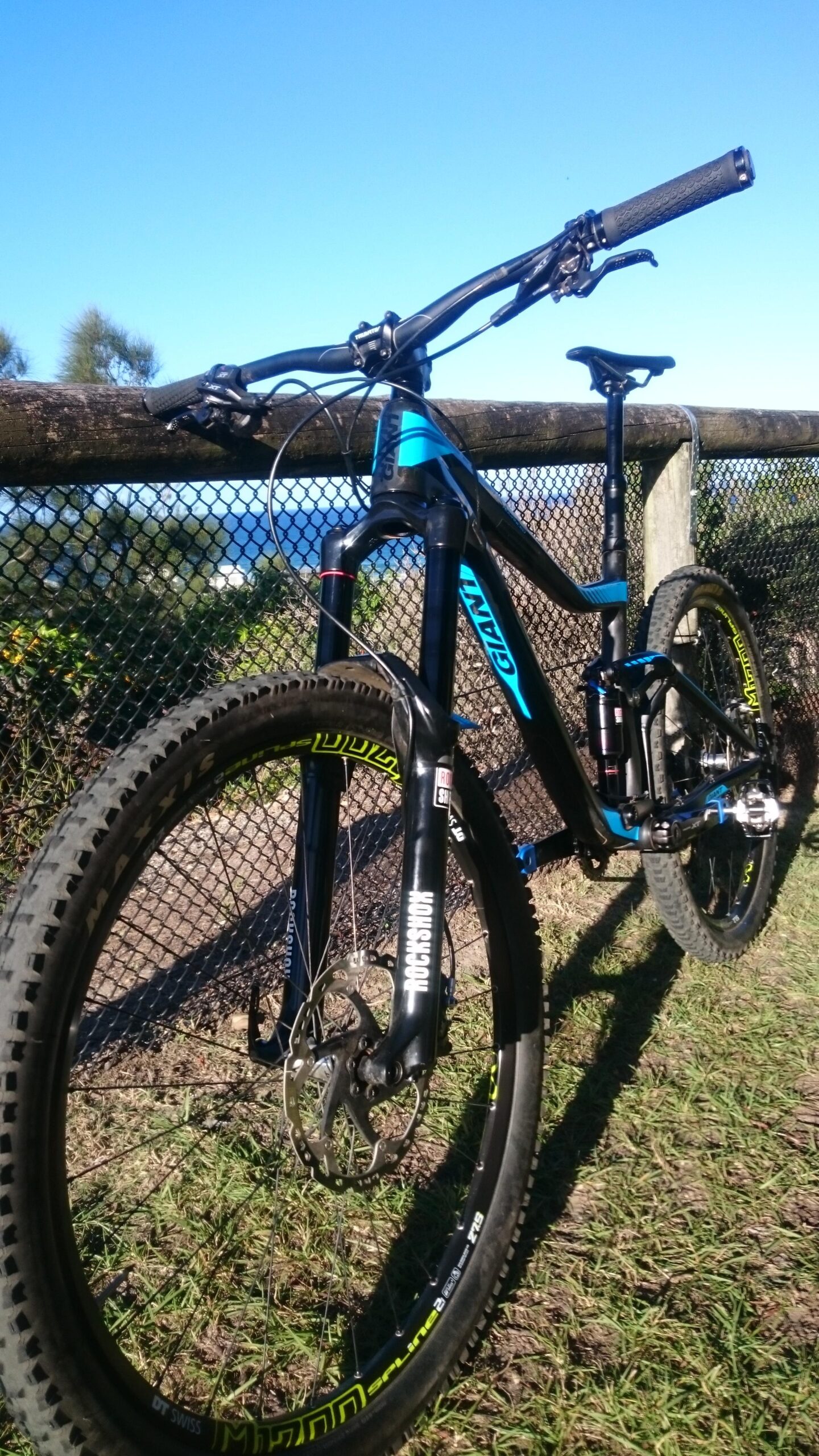 Giant Trance: A close-up view of a mountain bike leaning against a fence, showcasing its blue and black frame, prominent front suspension, and detailed tires with yellow accents. The background features green grass and a clear blue sky, suggesting an outdoor setting ideal for cycling.