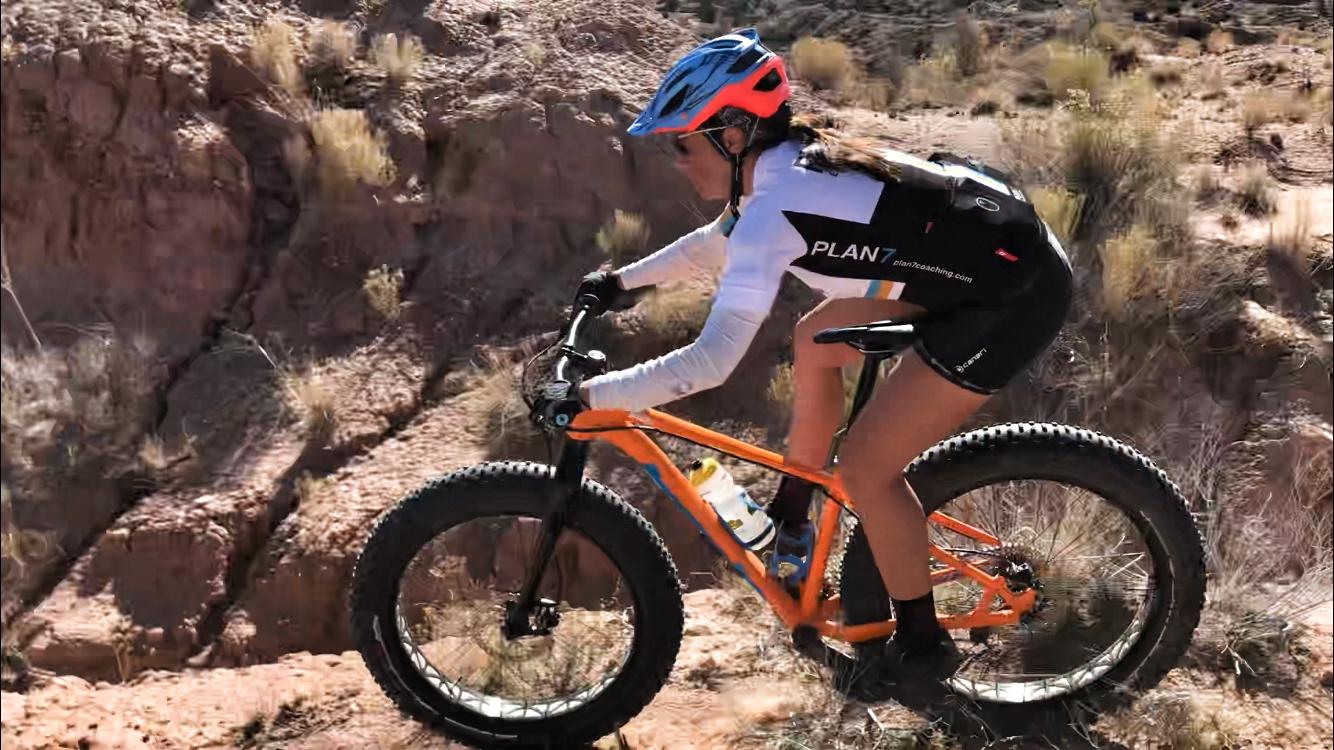 A person riding a bright orange fat bike on a rocky trail, wearing a blue and pink helmet, sunglasses, and a cycling kit with the logo "PLAN7." The terrain is arid, with sparse vegetation and steep rocky cliffs in the background. Mariposa Fat Bike Trails mountain bike trail.