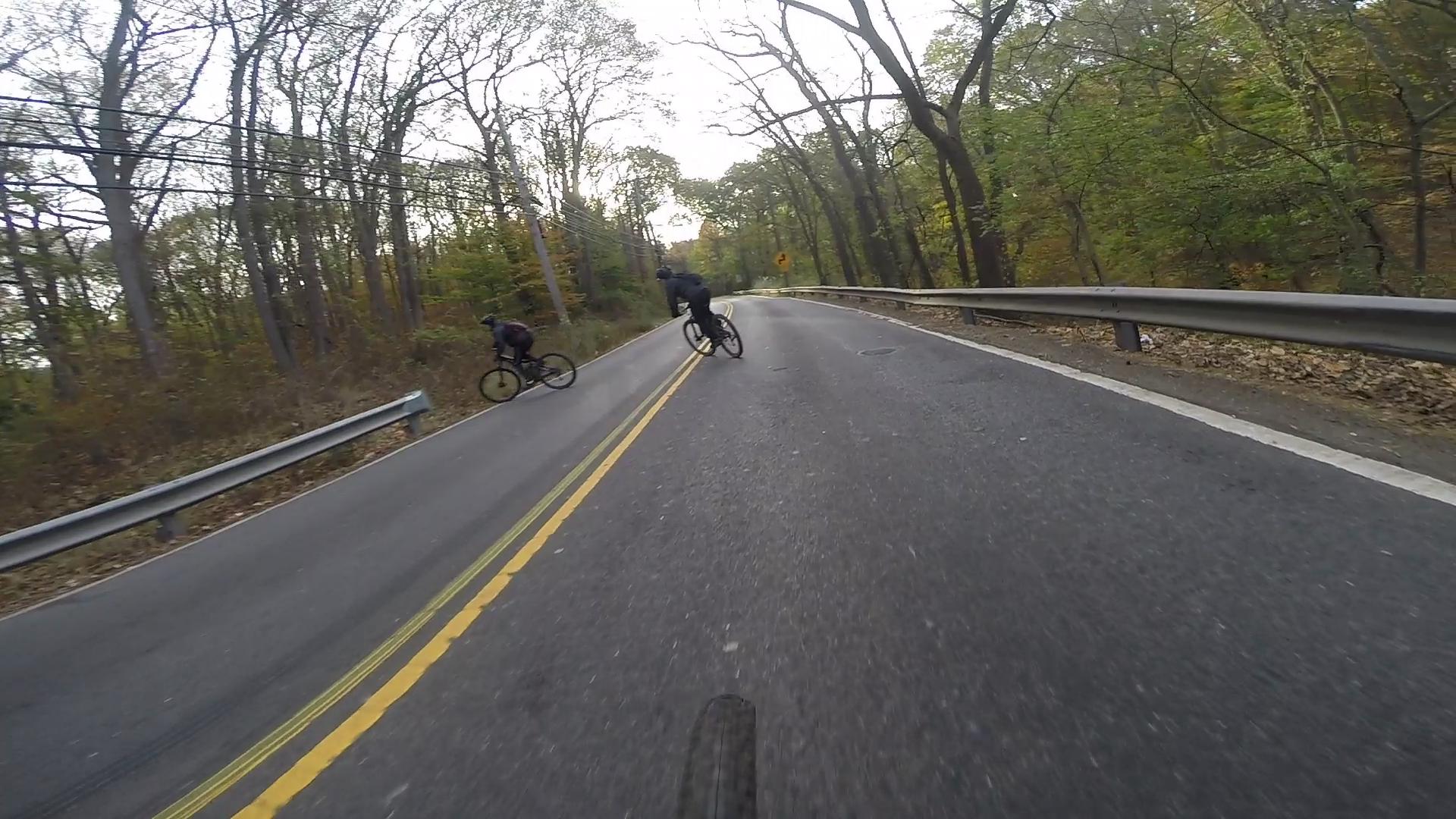 Two cyclists riding on a curved, two-lane road surrounded by trees in an autumn setting. One cyclist is in the foreground, positioned slightly to the left, while the other cyclist is further back on the right, both in motion.  Richmond Avenue and Forest Hill road mountain bike trail.