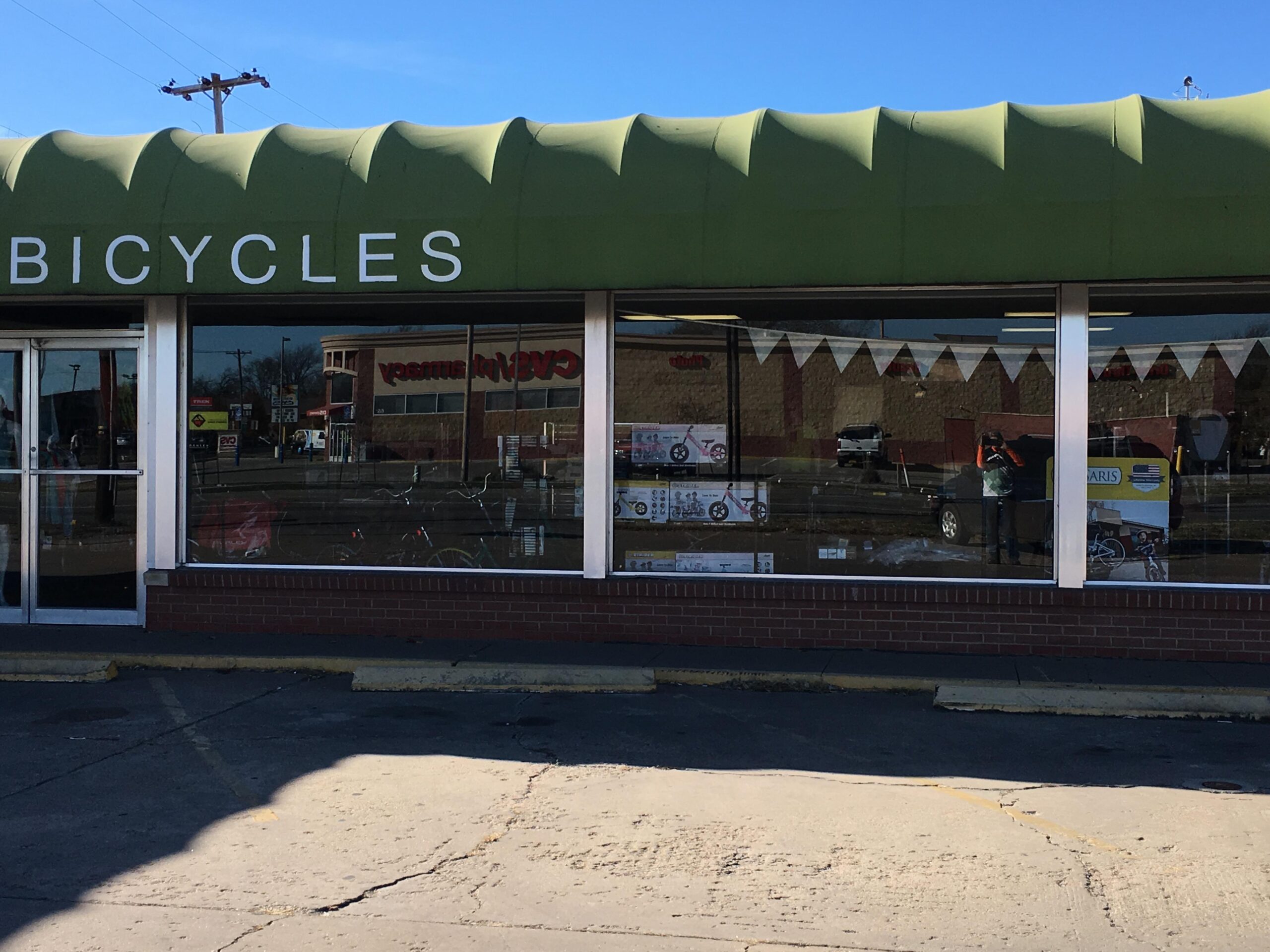 A bicycle shop with a green awning labeled "BICYCLES" is shown. The storefront features large windows displaying various bicycles and accessories inside. In the background, a sign for "CVS Pharmacy" is partially visible, along with other businesses. The scene is set on a clear day with a blue sky.