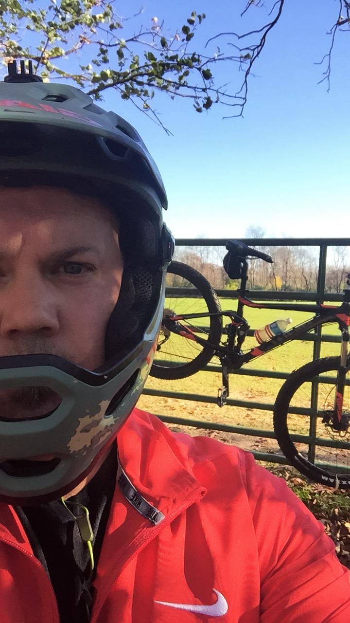 Giant Stance 27.5 2: A person wearing a helmet and a red jacket takes a selfie in front of a bike leaning against a fence, with a clear blue sky and trees in the background.