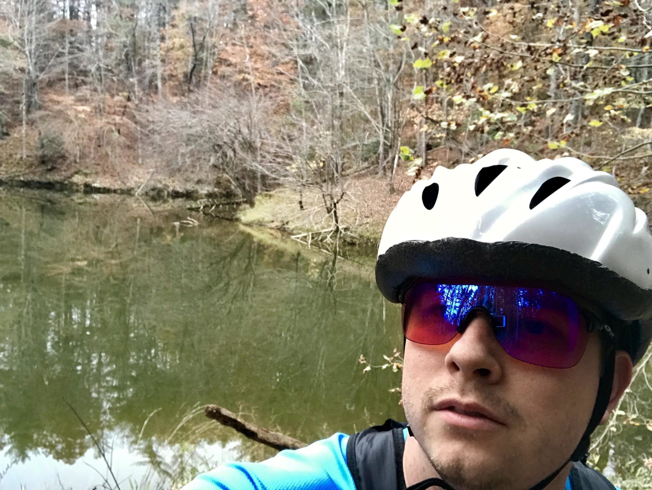 A person wearing a white biking helmet and sunglasses stands by a calm, reflective pond surrounded by bare trees and autumn foliage. Warrior Creek mountain bike trail.