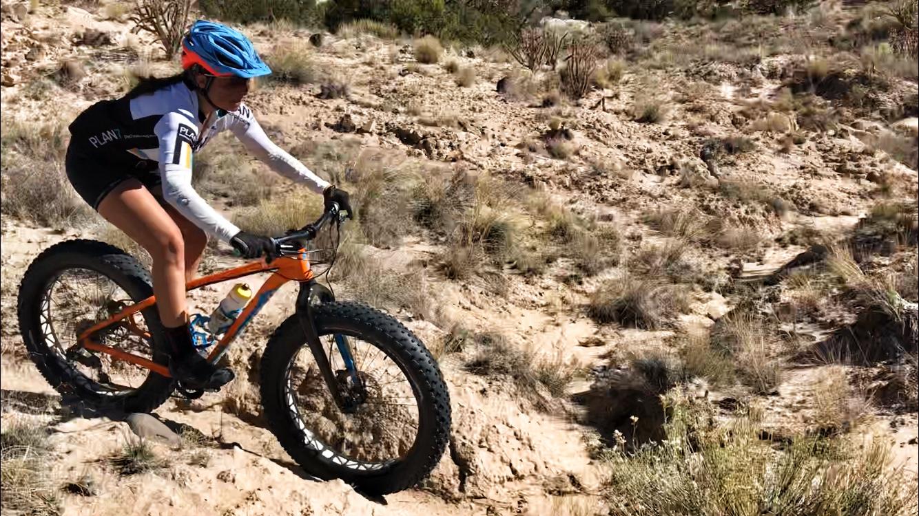 A person riding a fat bike on a rugged dirt trail, surrounded by sparse vegetation and rocky terrain under bright sunlight. The rider is wearing a helmet and athletic clothing, focusing on navigating the uneven landscape. Mariposa Fat Bike Trails mountain bike trail.