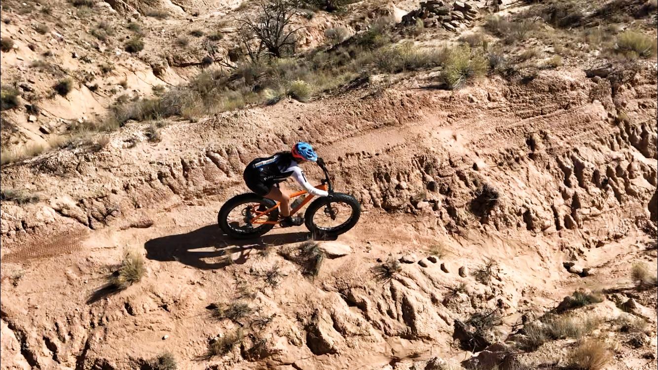 A cyclist riding a fat tire bike along a rugged, sandy trail in a desert landscape. The terrain features rocky outcrops and sparse vegetation, with the cyclist wearing a helmet and athletic gear, navigating a steep incline. Mariposa Fat Bike Trails mountain bike trail.