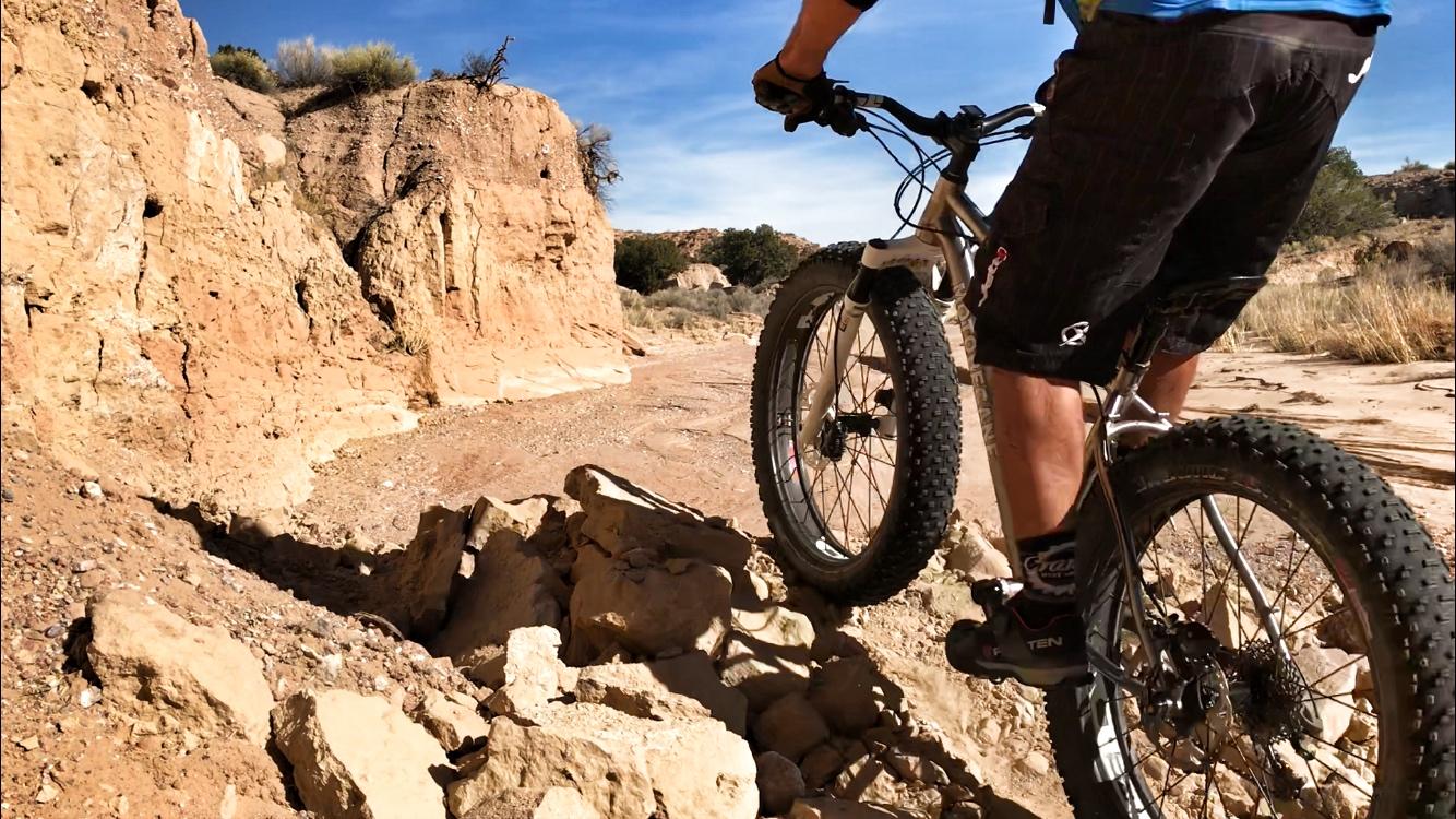 A person riding a fat tire bicycle on rocky terrain, maneuvering over boulders, with a clear blue sky and desert landscape in the background. Mariposa Fat Bike Trails mountain bike trail.