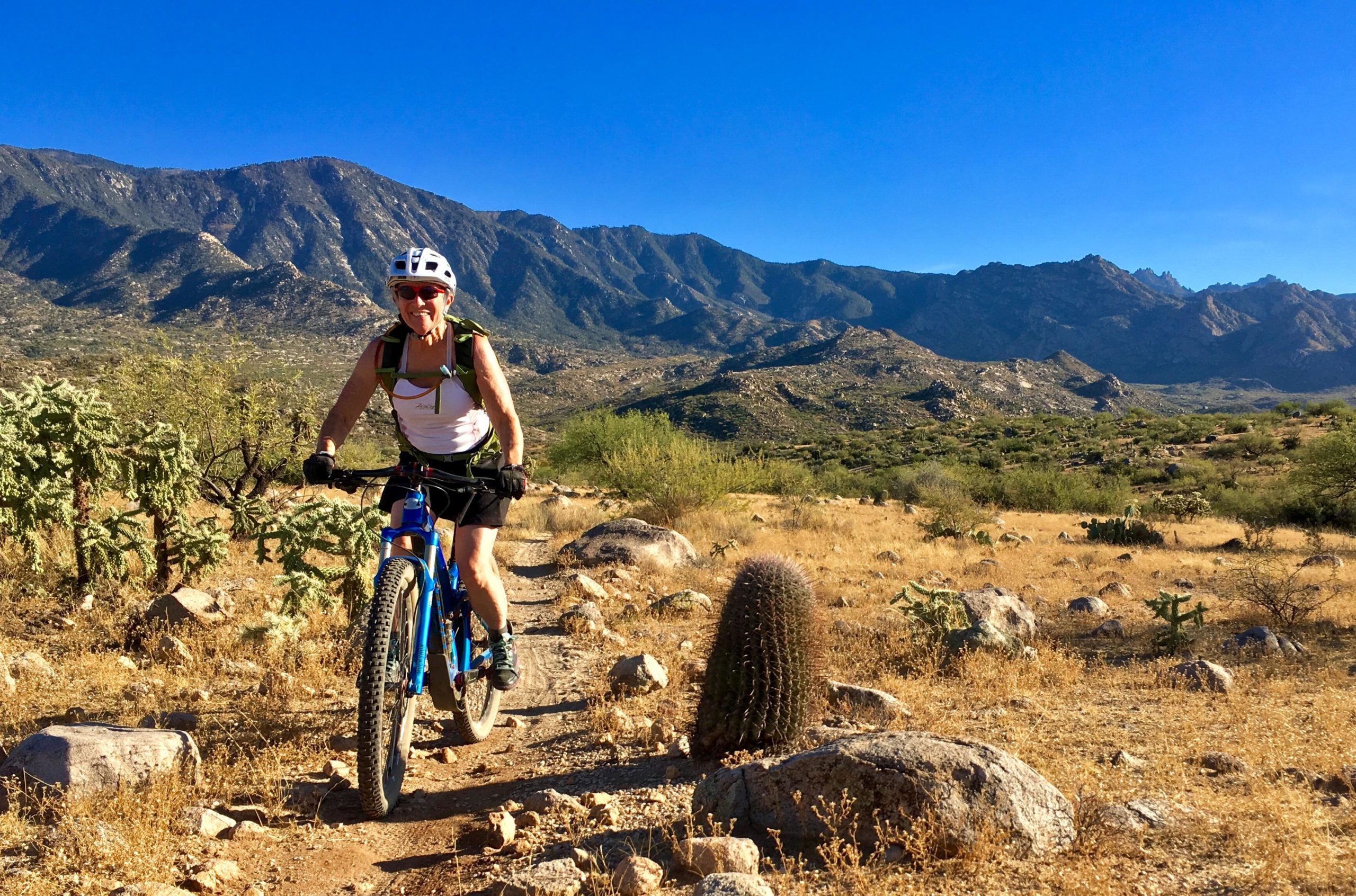 A person riding a blue mountain bike on a gravel trail surrounded by a desert landscape. In the background, there are mountains under a clear blue sky, and the foreground includes cacti and rocky terrain. The rider is wearing a helmet and sunglasses, smiling as they navigate the trail. 50-year Trail / Golder Ranch mountain bike trail.