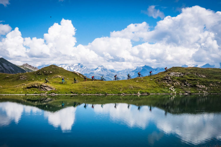 A scenic view of a mountain landscape featuring cyclists riding along a grassy ridge beside a serene lake. The calm water reflects the clouds and surrounding mountains under a bright blue sky.