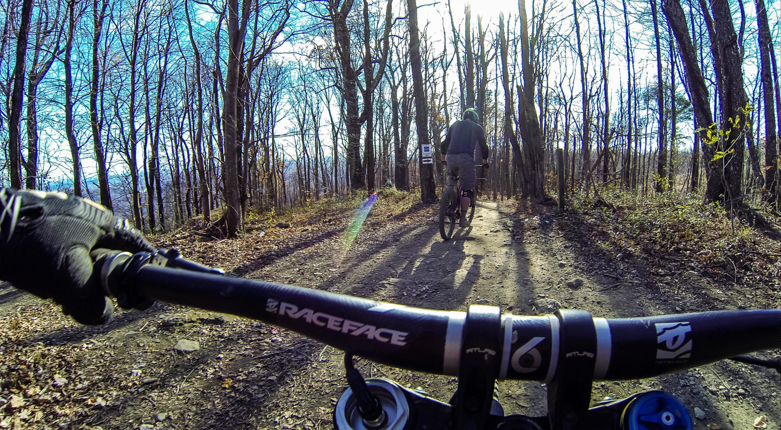 A view from the handlebars of a mountain bike, showing a dirt trail winding through a wooded area with bare trees. In the background, another cyclist is riding ahead, and sunlight filters through the branches, creating a bright atmosphere. Windrock Bike Park mountain bike trail.