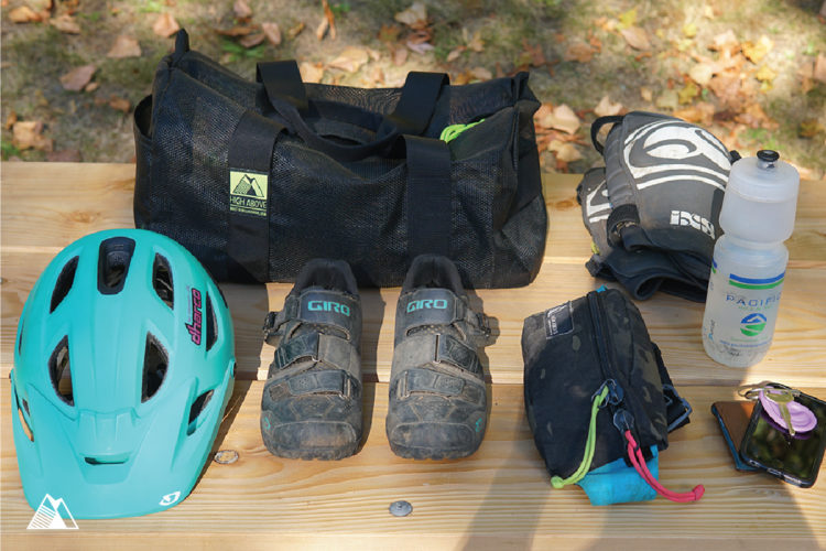 A selection of cycling gear laid out on a wooden surface, including a teal helmet, black cycling shoes, a black duffel bag, a water bottle, a pair of gloves, a small zippered pouch, and a smartphone. Leafy surroundings suggest an outdoor setting.