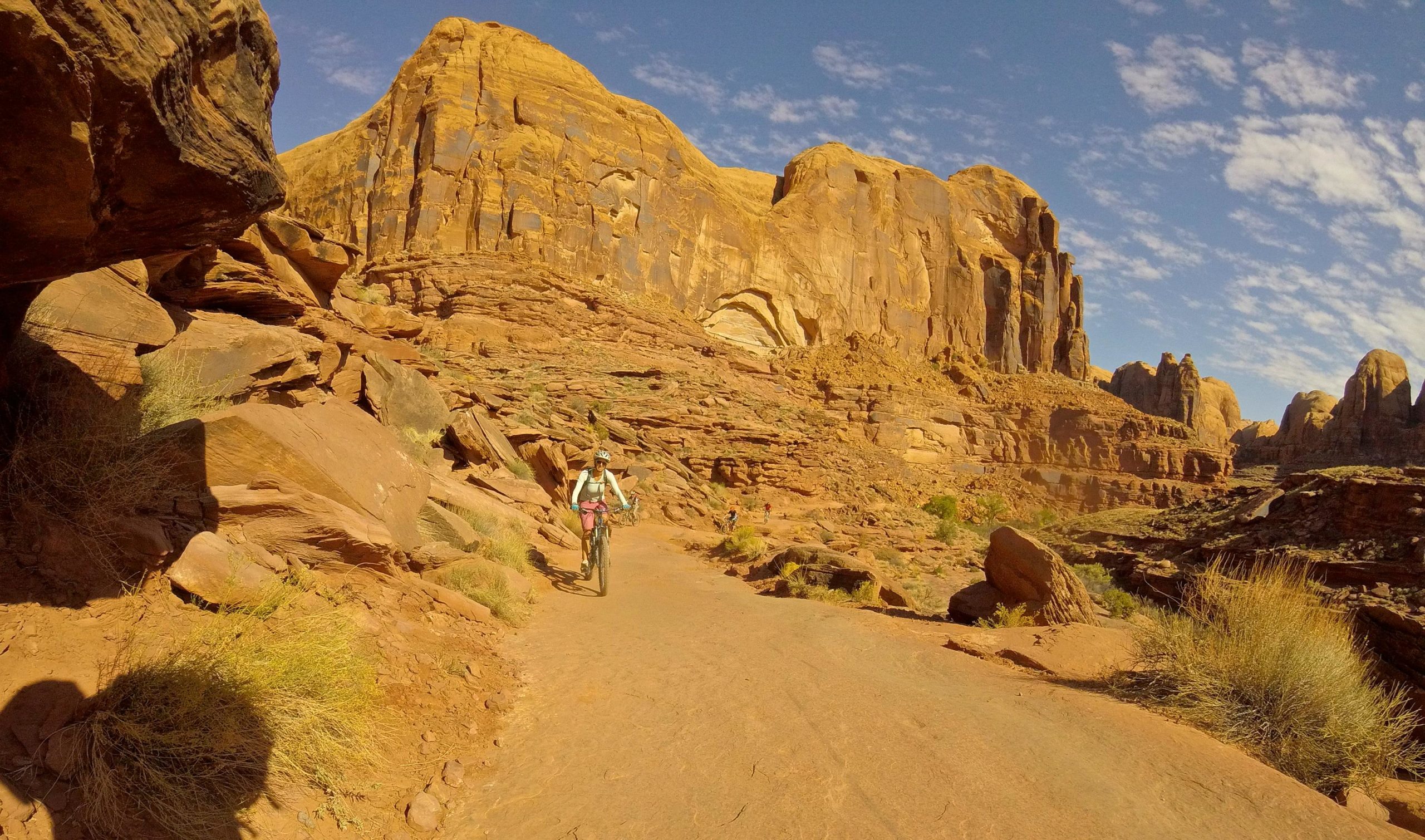 A mountain biker rides along a dirt path in a rugged desert landscape, surrounded by towering red rock formations and scattered vegetation under a partly cloudy blue sky. Hymasa mountain bike trail.