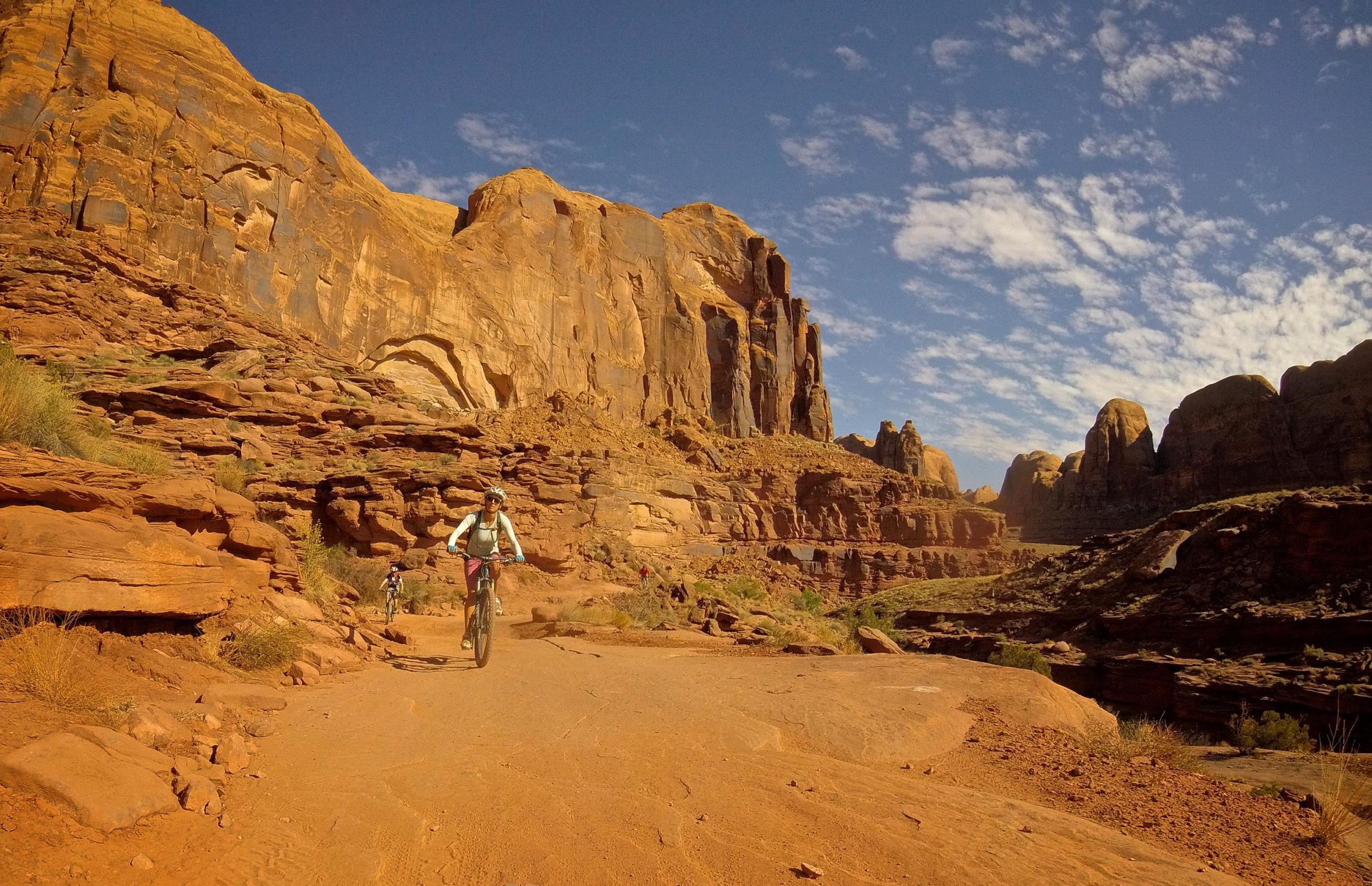 A mountain biker rides along a dusty trail surrounded by towering red rock formations under a blue sky with clouds. The rugged landscape features steep cliffs and sparse vegetation, creating a scenic backdrop for outdoor adventure. Hymasa mountain bike trail.