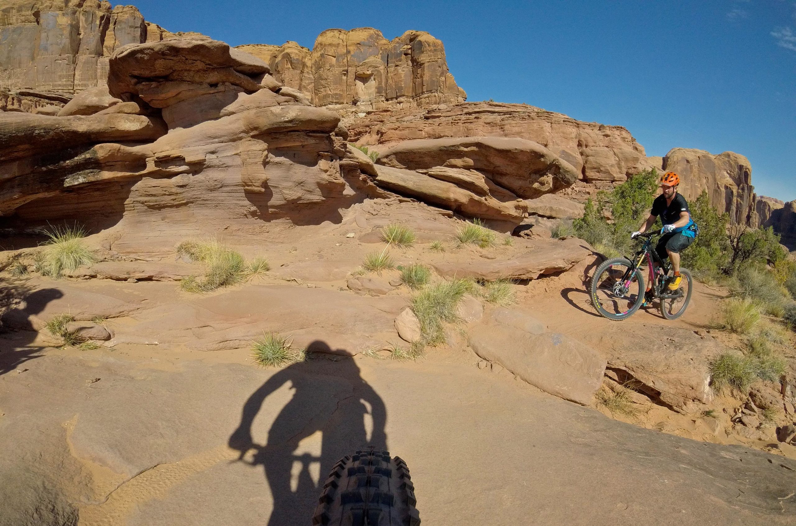 A person riding a mountain bike on rocky terrain with tall cliffs in the background, casting a shadow on the ground. The scene features grasses and shrubs amidst the stone landscape under a clear blue sky. Hymasa mountain bike trail.