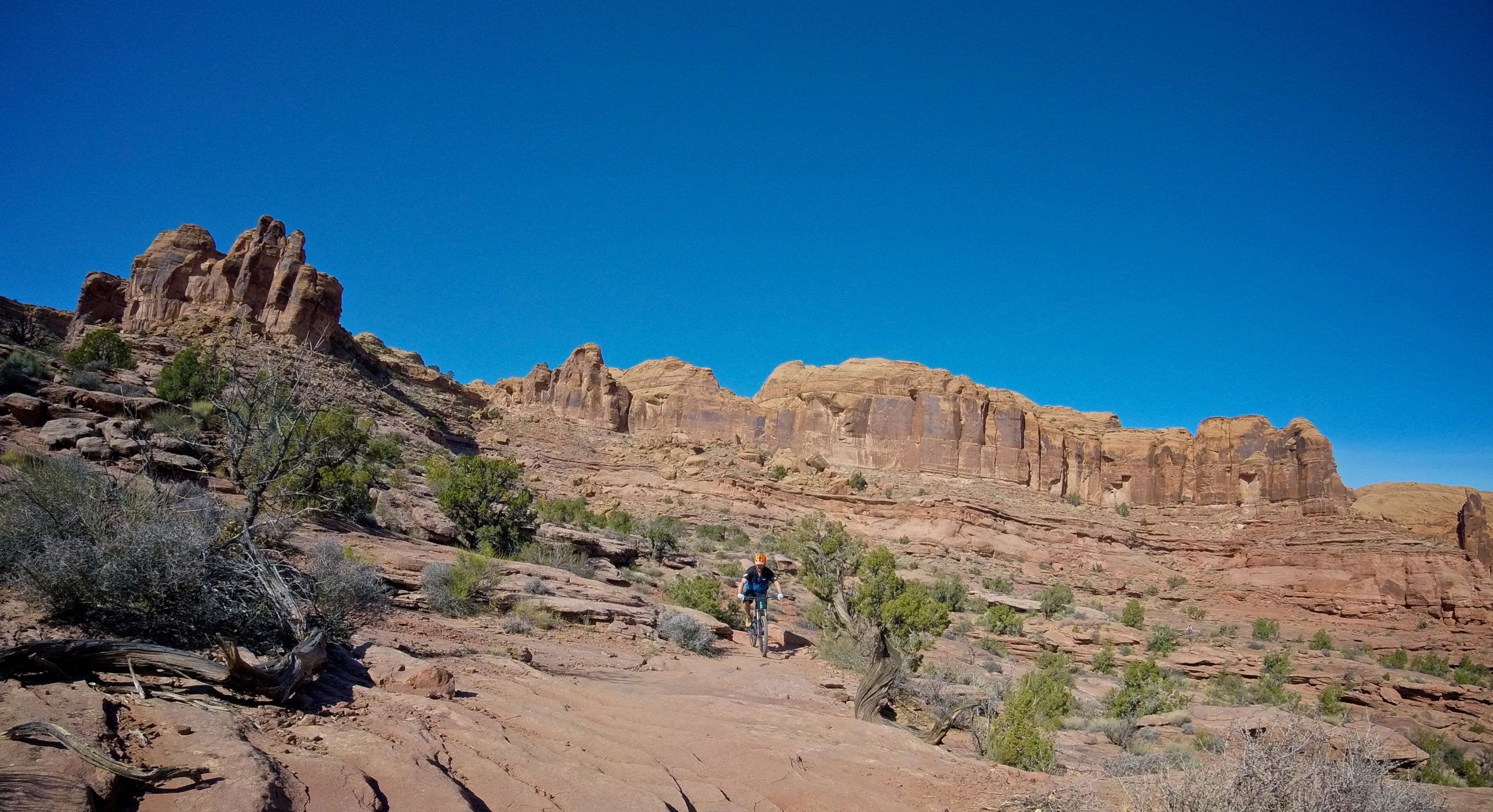 A mountain biker navigating a rocky trail in a desert landscape, surrounded by red rock formations and sparse vegetation under a clear blue sky. Hymasa mountain bike trail.