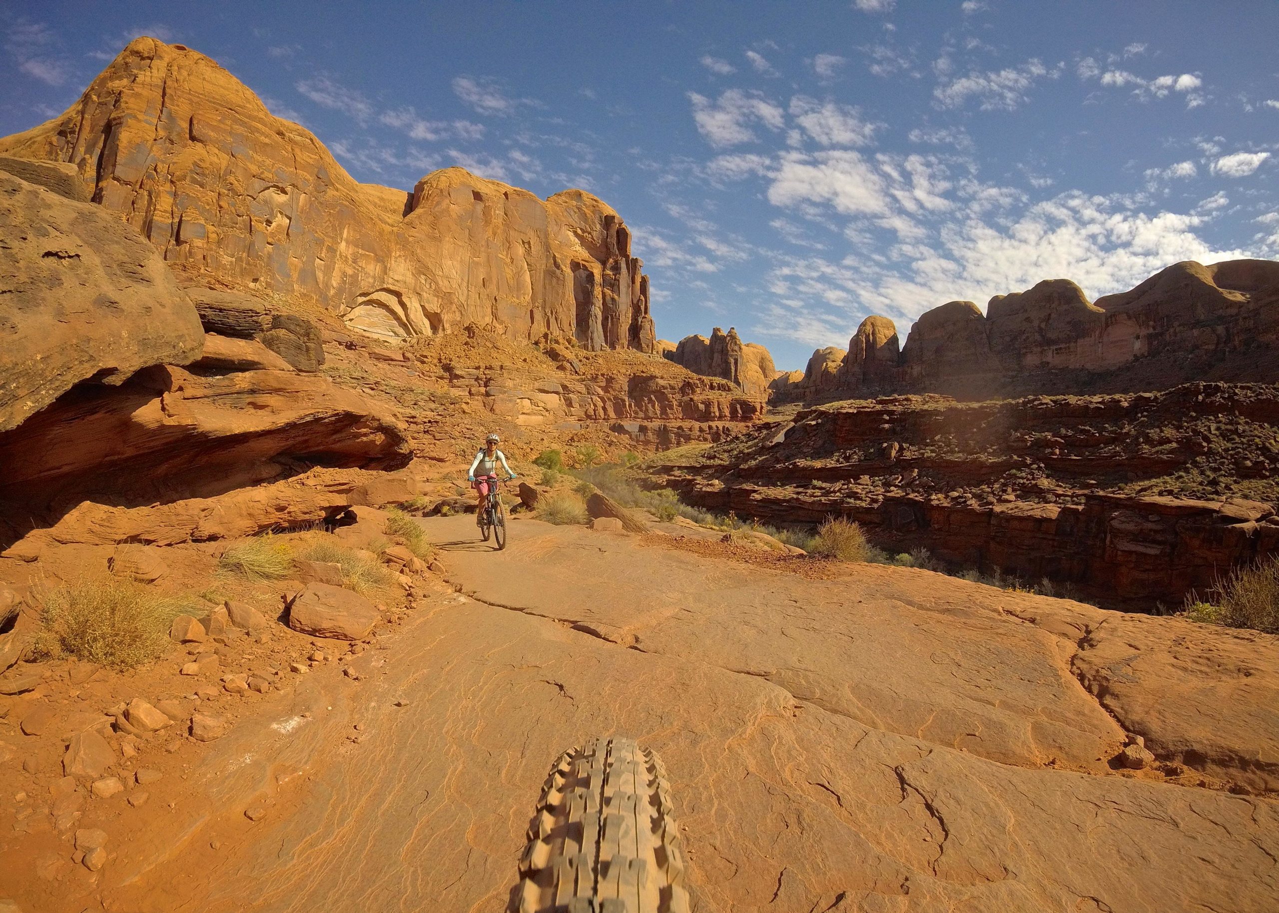 A mountain biker riding along a rugged dirt trail in a desert landscape, with towering red rock formations and a blue sky filled with scattered clouds in the background. Hymasa mountain bike trail.