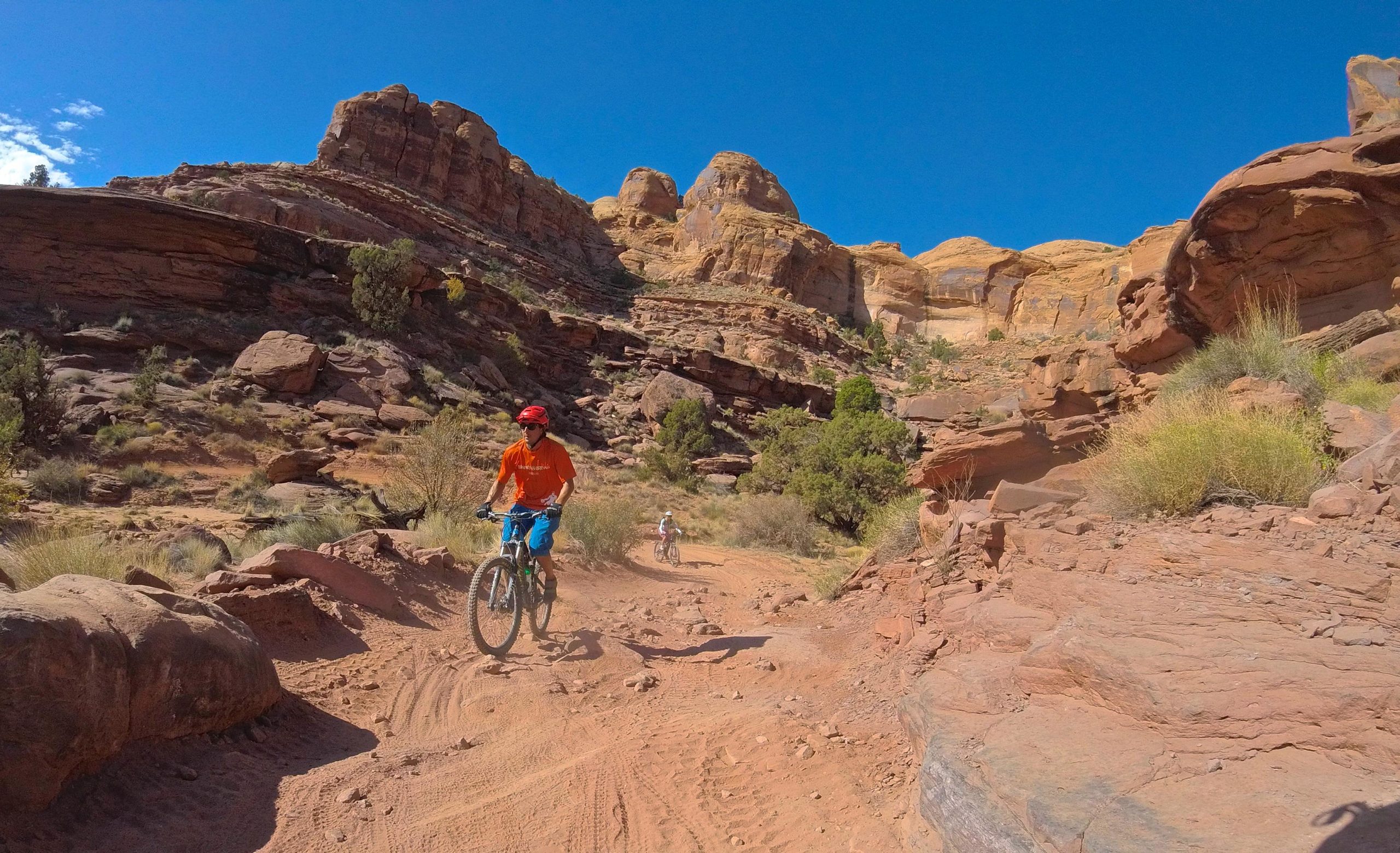 Two mountain bikers riding on a dirt trail through a rocky canyon landscape under a clear blue sky. The trail is surrounded by red rock formations and sparse vegetation. One biker is wearing an orange shirt and a red helmet, while the other is further back on the trail. Hymasa mountain bike trail.