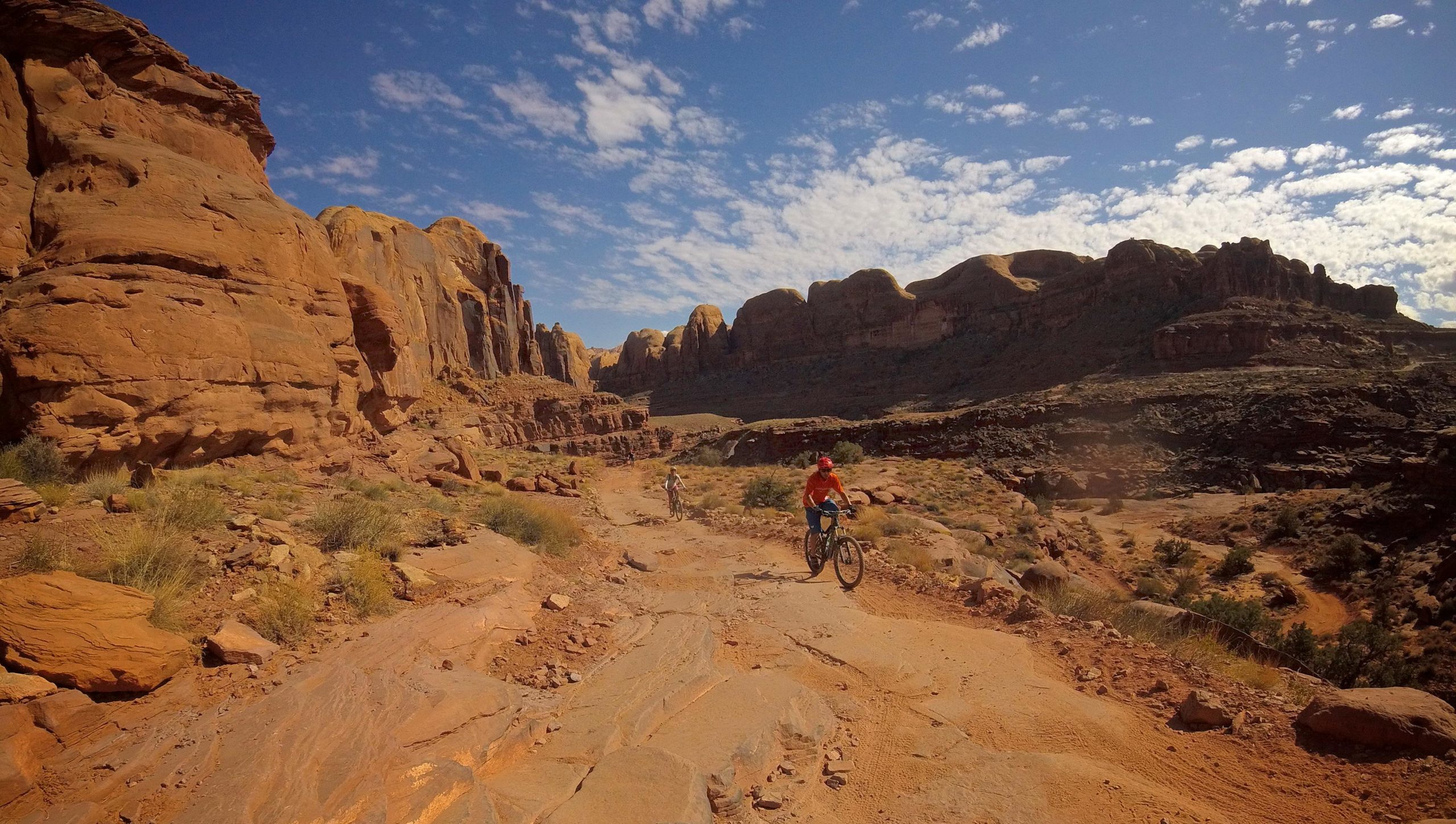 Two mountain bikers navigate a rocky dirt trail surrounded by striking red rock formations and a clear blue sky with scattered clouds. The landscape features rugged terrain, shrubs, and towering cliffs, conveying an adventurous outdoor setting. Hymasa mountain bike trail.