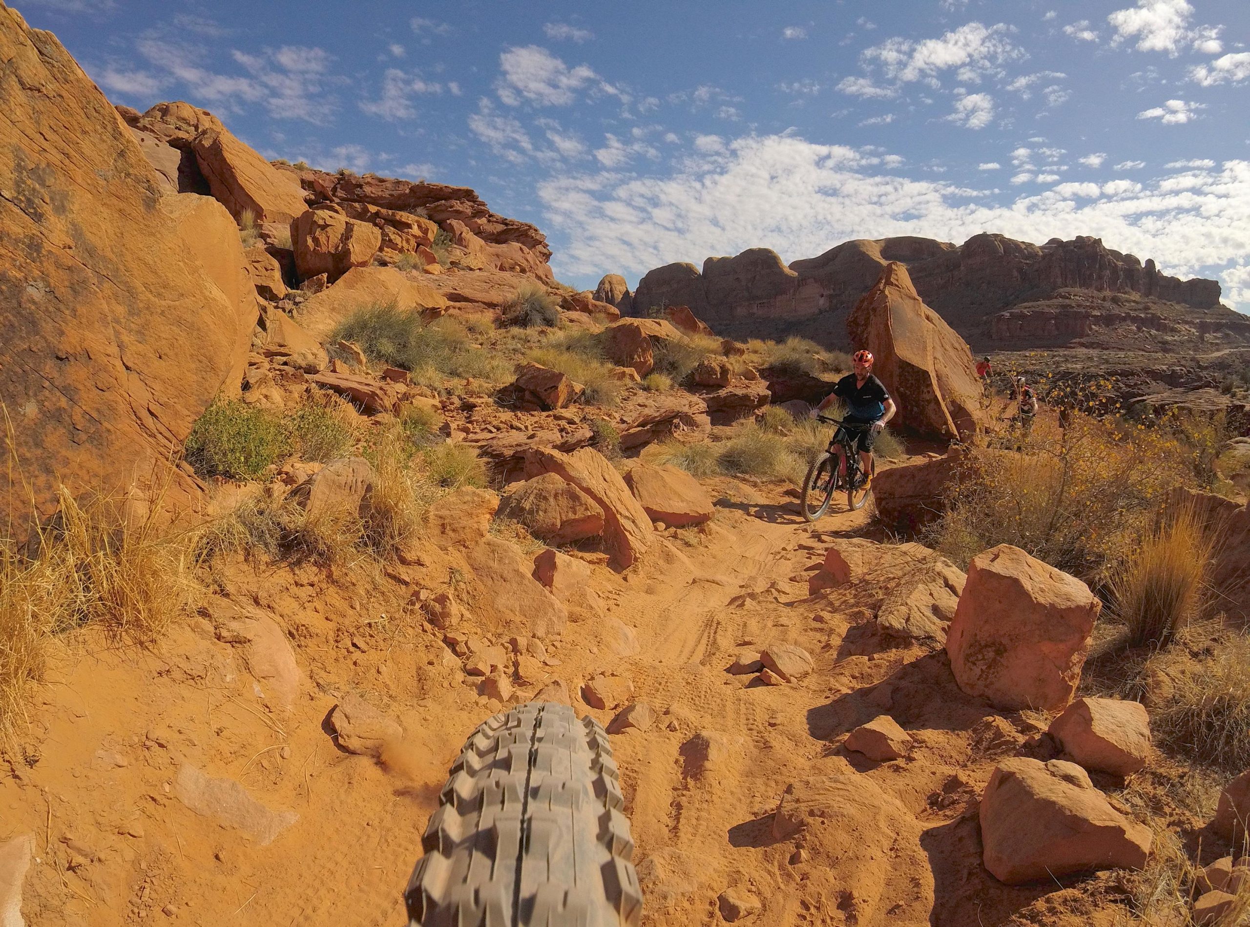 A mountain biker navigating a rocky desert trail, with bright orange-red rocks and sparse vegetation around. The scene features a blue sky with scattered clouds in the background and showcases the rugged terrain typical of outdoor biking adventures. Hymasa mountain bike trail.