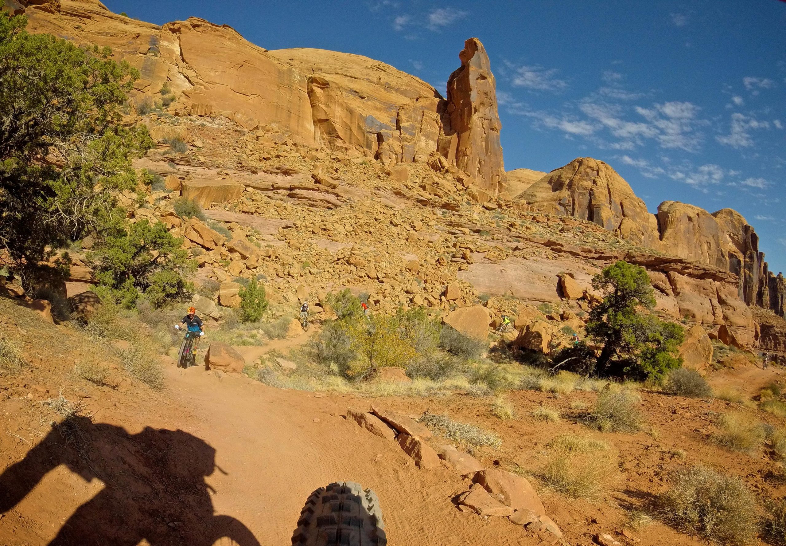 Mountain bikers navigate a rocky trail surrounded by striking red rock formations and desert vegetation under a clear blue sky. The image captures a dynamic outdoor adventure scene. Hymasa mountain bike trail.