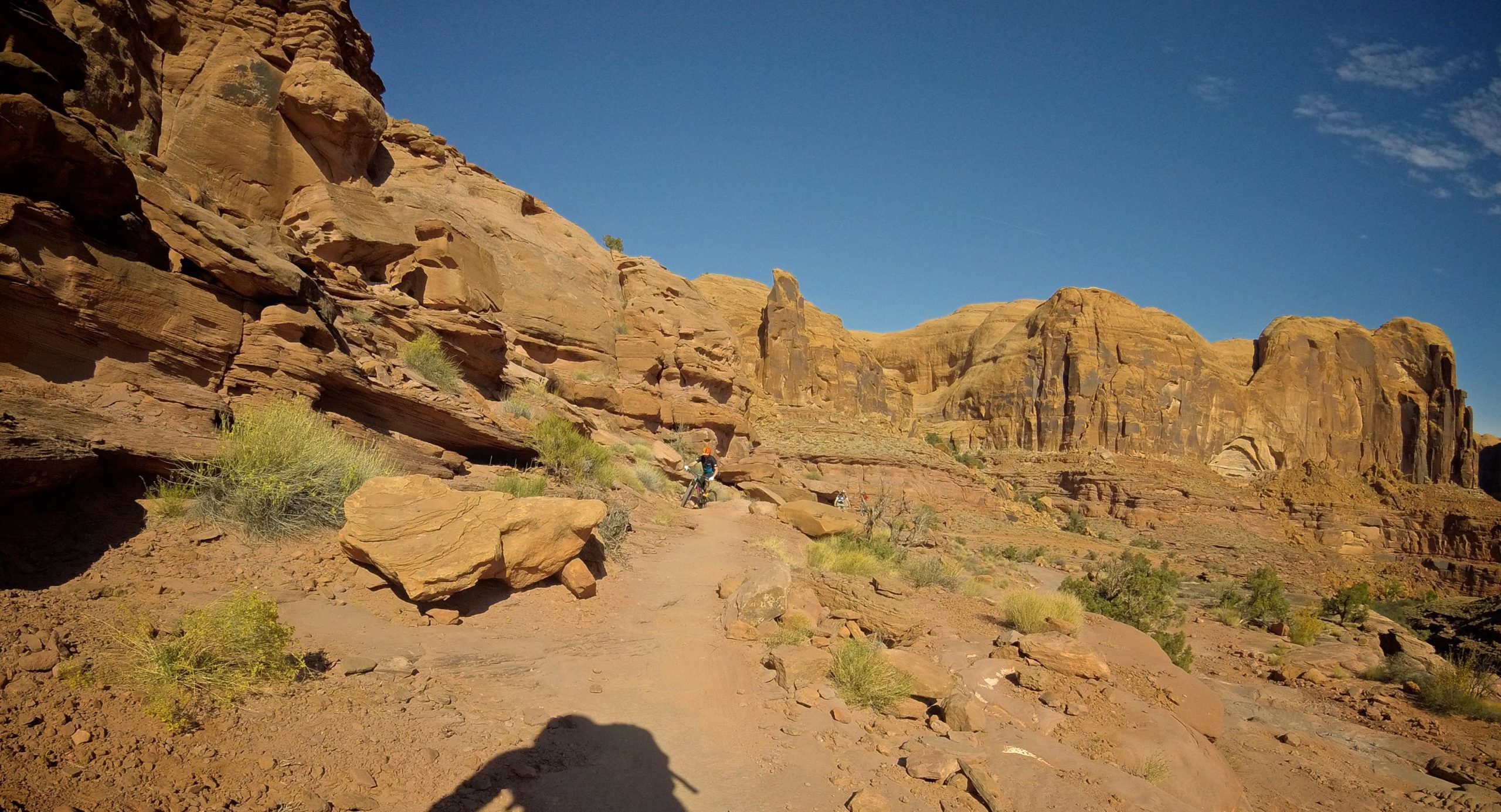 A winding dirt path leads through a dramatic landscape of red rock formations and cliffs under a clear blue sky. The terrain features scattered vegetation and shadows cast by the rugged cliffs, creating a picturesque scene of outdoor adventure. Hymasa mountain bike trail.