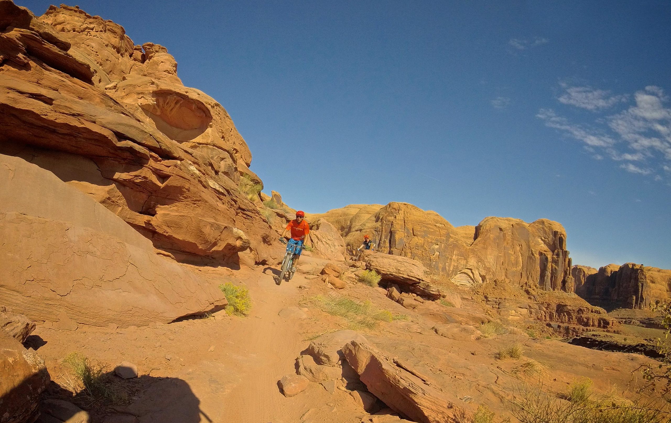 A mountain biker navigates a rocky trail in a desert landscape, surrounded by large red rock formations under a clear blue sky. Vegetation appears sparse on the sandy ground. Hymasa mountain bike trail.
