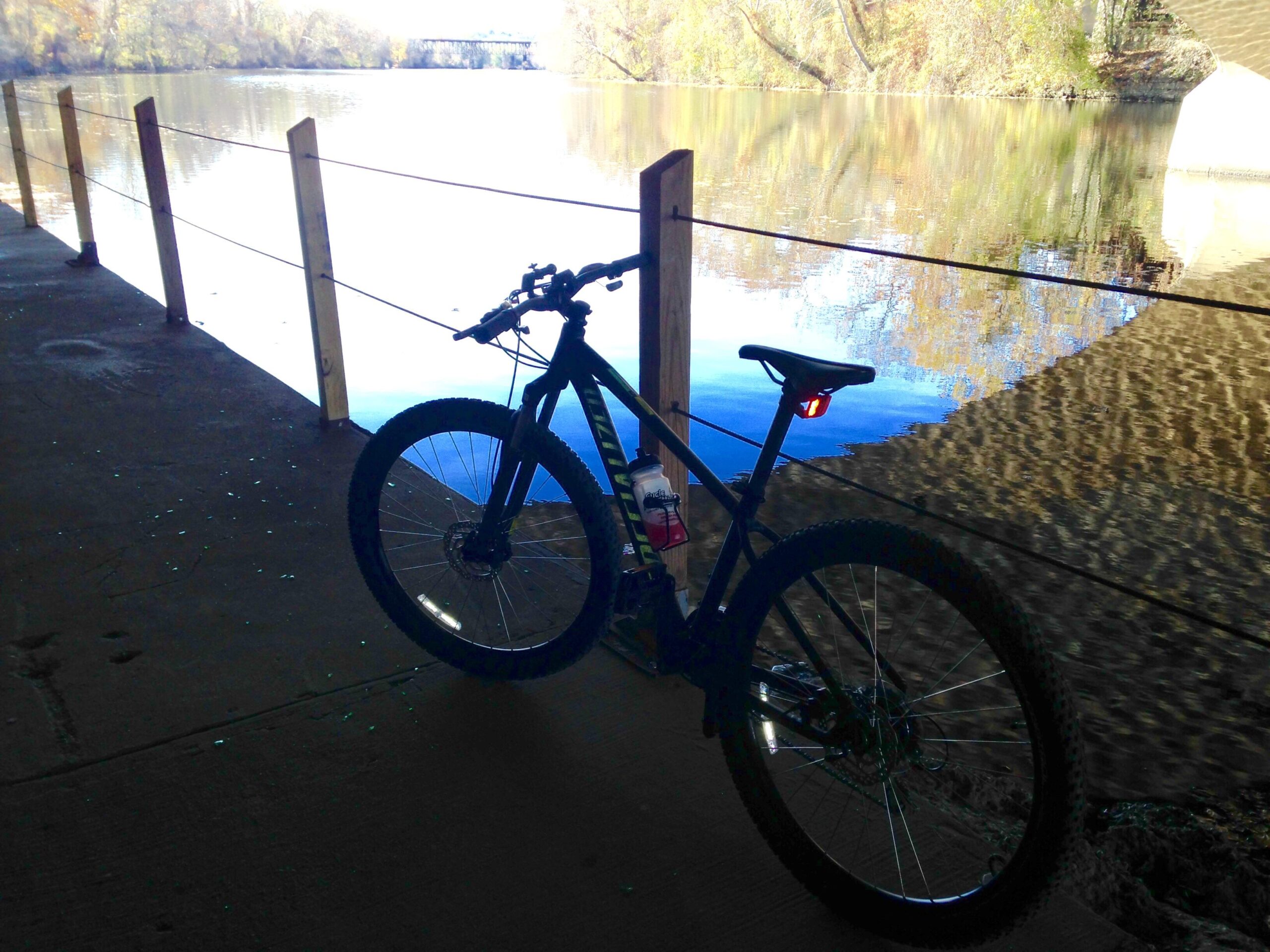 Specialized Pitch Sport 650B: A mountain bike leaning against a railing under a bridge, with a calm river reflecting the surrounding trees and light. The scene is peaceful and showcases the natural beauty of the area.