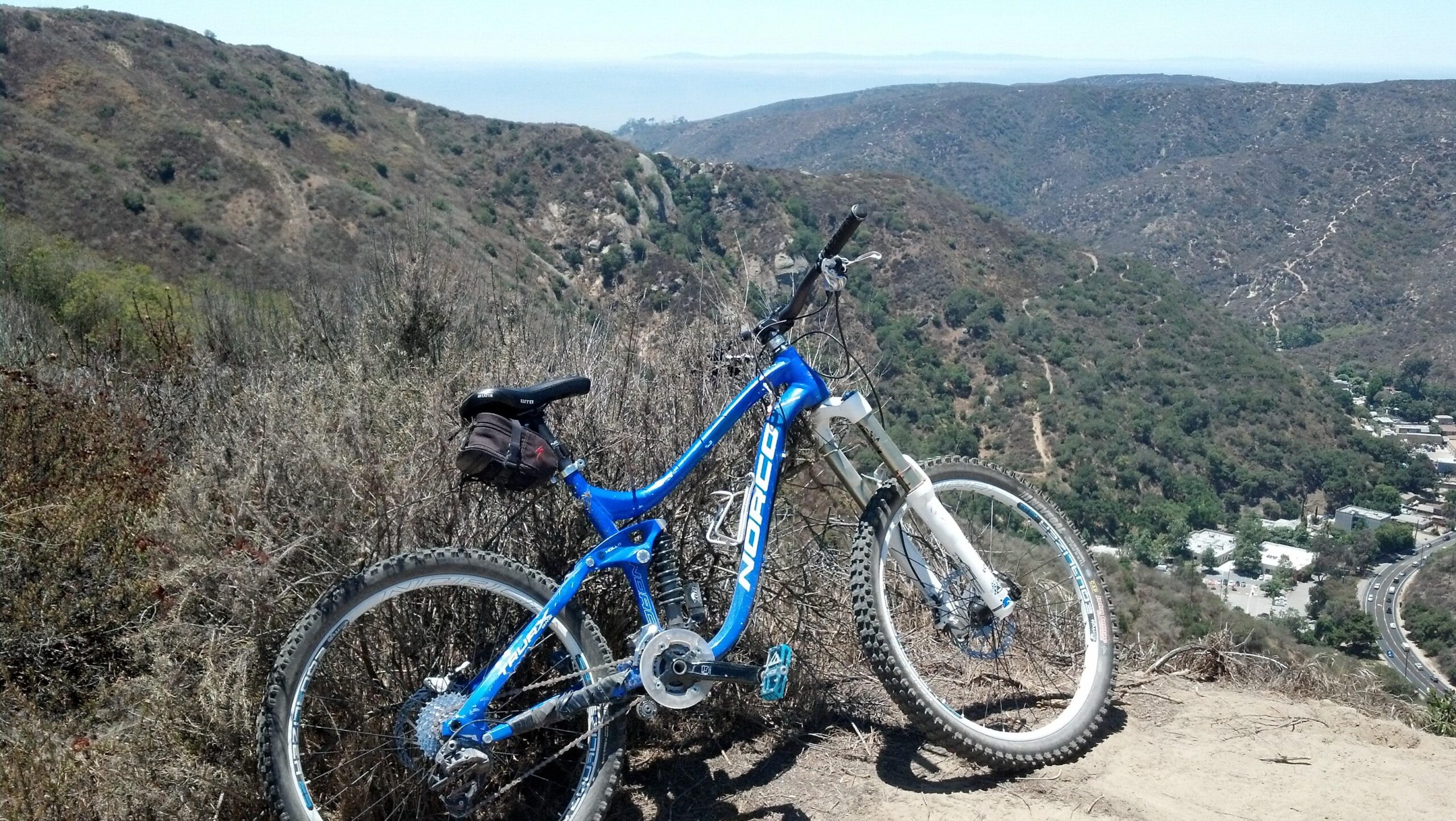 Norco TRUAX 1: A blue mountain bike is resting on a dirt path overlooking a hilly landscape. The background features rolling hills covered with greenery, and a glimpse of the ocean is visible in the distance beneath a clear sky. The scene captures a moment of tranquility in nature, ideal for outdoor enthusiasts.